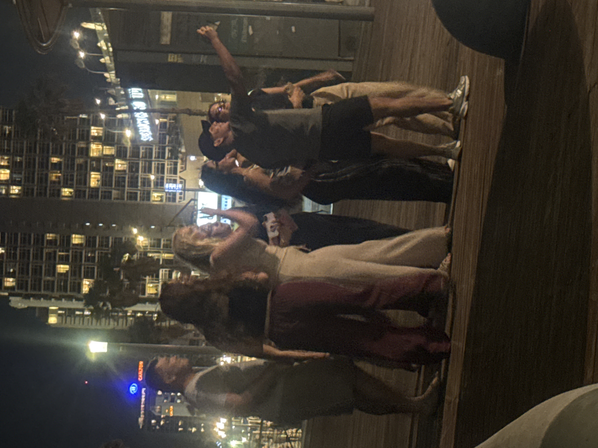 The image captures a nighttime scene on a wooden promenade in Tel Aviv, Israel, featuring a group of approximately eight individuals. In the background, multiple high-rise buildings with numerous illuminated windows define the urban skyline, interspersed with palm trees. A prominent blue-lit sign displaying "CARLTON" is visible on a building in the mid-left background. To the right, another illuminated sign in Hebrew clearly reads "שדרות ירושלים" (Jerusalem Boulevard).
In the immediate foreground, a group of people is engaged in a social interaction. On the far right, a man wearing a dark t-shirt, dark shorts, and white sneakers extends his right arm, holding a smartphone outward, indicative of taking a selfie or group photograph. To his left, a woman in a dark top and light-colored pants, and a man wearing a dark baseball cap, dark t-shirt, and dark shorts, are oriented towards the smartphone. Further left, a woman in a dark top and wide, burgundy-colored pants stands next to a woman in a light-colored top and light wide-leg pants. On the far left, a man in a light t-shirt and shorts is partially visible, facing towards the right of the frame. The wooden promenade surface is visible in the lower half of the image. Artificial streetlights illuminate the scene, creating areas of brightness and shadow across the subjects and environment.