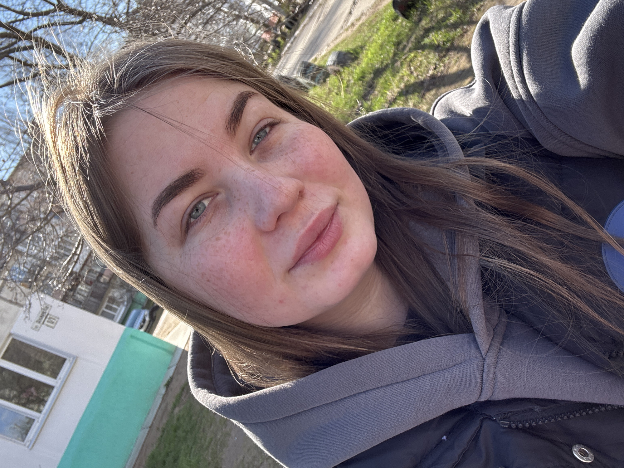A young woman is featured in a close-up outdoor selfie, likely taken in Chaikovsky, Russia. She has long, straight brown hair, bright green eyes, and visible freckles on her cheeks and nose. She wears a grey hooded sweatshirt layered over a darker, possibly padded jacket. Her expression is soft and natural, with a subtle smile.
The setting is outdoors on a bright, sunny day, indicating clear weather. The time of day appears to be during daylight hours. In the background, bare tree branches are visible against a light blue sky, suggesting late winter or early spring. A building is present in the left background; it's light-colored with white-framed windows and a distinct bright green base. Patches of green grass can be seen on the ground, further indicating the season.
A notable detail on the building is a rectangular sign with Cyrillic text. Portions of the text, visible in the crops, show "Воробьева" (Vorobyova), which could be a street name or a surname, and possibly a building number, like "б. 1". The sunlight illuminates the woman's hair, giving it a highlighted appearance. No specific activity beyond posing for the selfie is depicted.
