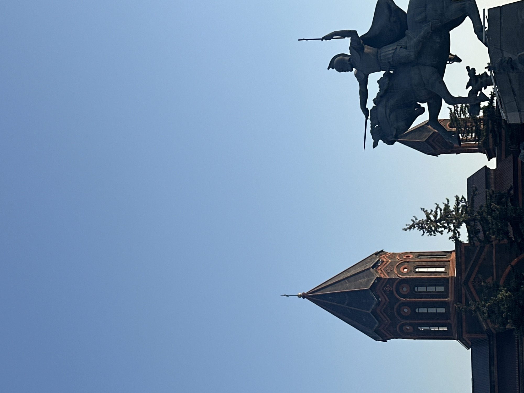 The image depicts a monument of a man on horseback in the foreground, with a church in the background. The monument is a dark, silhouetted figure of a warrior wearing a helmet and cape, holding a sword and a staff, with the horse in mid-stride. The church features a conical, dark-colored roof with intricate red and black patterns and arched windows. Several evergreen trees are visible between the monument and the church. The sky is clear and a bright blue. The monument is identified as the statue of Vardan Mamikonian, a key figure in Armenian history, and the church is the Holy Mother of God Cathedral in Gyumri, Armenia. The scene is captured from a low angle, emphasizing the grandeur of the monument and the cathedral against the expansive sky.