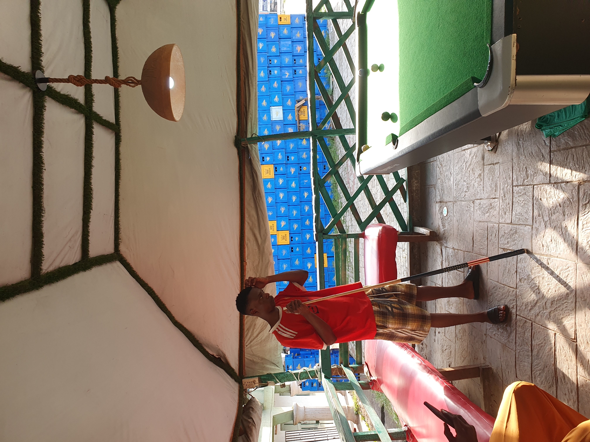 In Lokoja, Nigeria, a young man stands near a pool table with a cue stick in hand. He is wearing a red t-shirt with white stripes on the sleeves and plaid shorts. The pool table is covered in green felt and has several balls on it. Behind the pool table is a fence made of green latticework. Beyond the fence, stacks of blue and yellow crates are visible, suggesting a storage or distribution area. The scene is set under a white, tent-like structure with some artificial grass detailing along the edges of the panels and a hanging lamp with a natural material shade. The ground is paved with stone tiles, and there is a red bench to the left of the pool table. The lighting suggests it is daytime, with shadows cast on the ground.