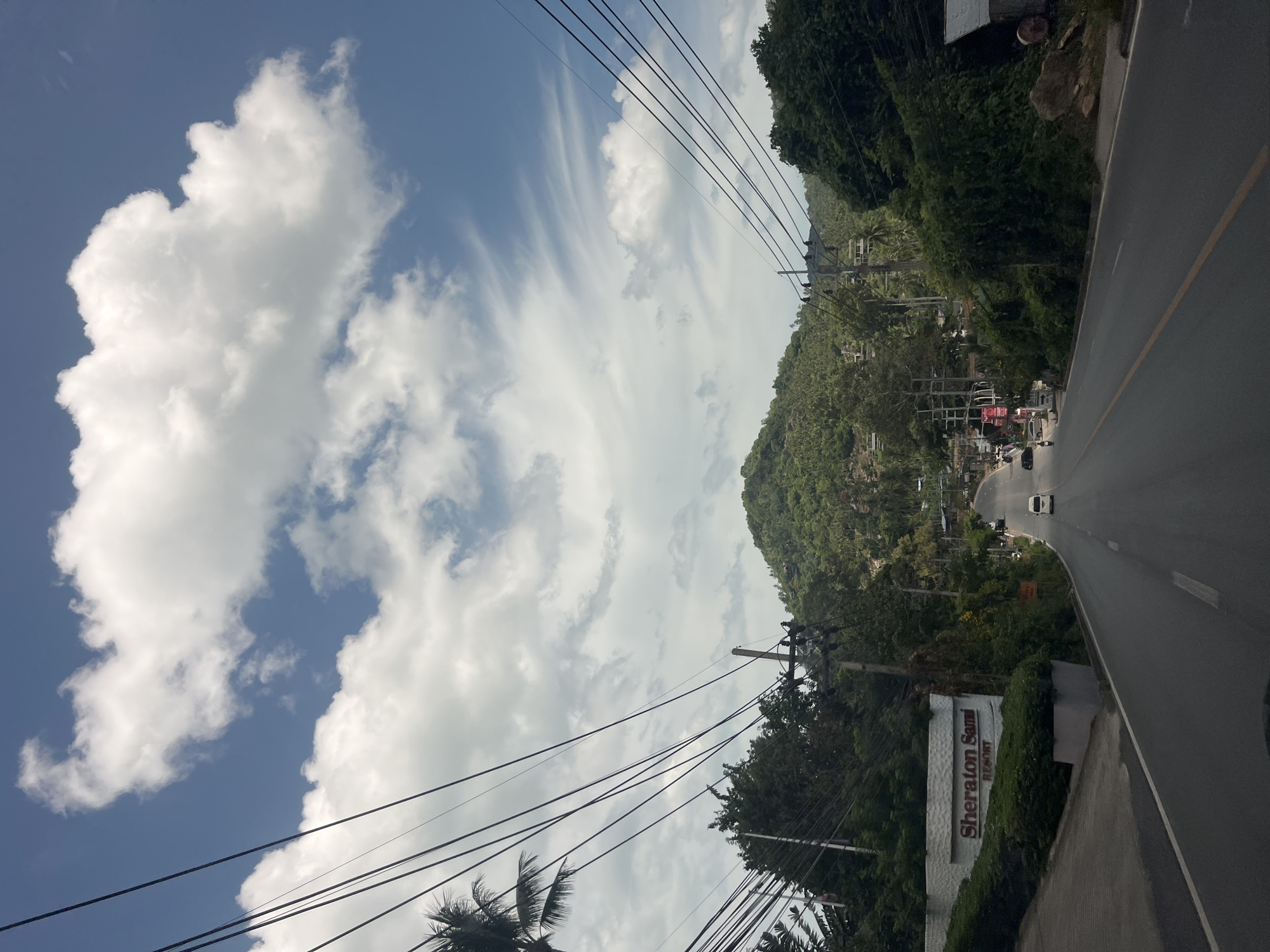 The image captures a scenic view of a road in Ko Samui District, Thailand, leading towards a lush, tree-covered mountain under a cloudy sky. On the left side of the road, a prominent sign for "Sheraton Samui Resort" is visible, bordered by manicured bushes. Overhead, a complex network of power lines crisscrosses the sky, supported by utility poles. The road itself is paved with a visible yellow line dividing the lanes. A few vehicles, including a white van and a motorcycle, are seen on the road. The surrounding environment is characterized by dense greenery, with palm trees and other tropical vegetation lining the sides of the road and climbing the slopes of the mountain. The overall impression is that of a tropical, well-traveled route in a resort area.