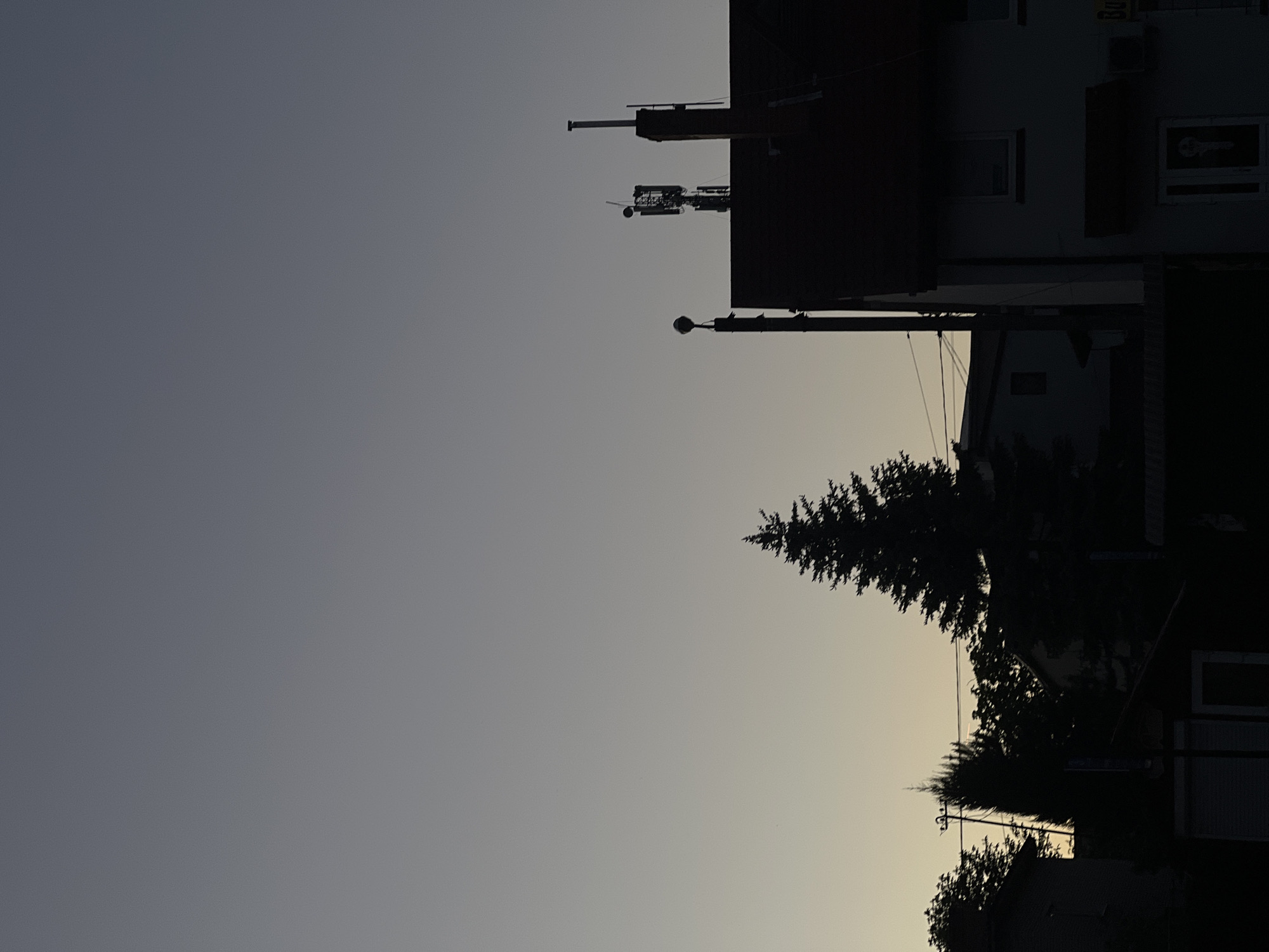 The image depicts a dusk scene in Horodok, Ukraine, featuring the silhouettes of buildings, trees, and utility poles against a gradient sky. A cell tower is visible atop one of the buildings. Trees line the lower right portion of the frame, obscuring the lower parts of the buildings. The sky transitions from a dark grey-blue above to a light yellowish-grey near the horizon.
