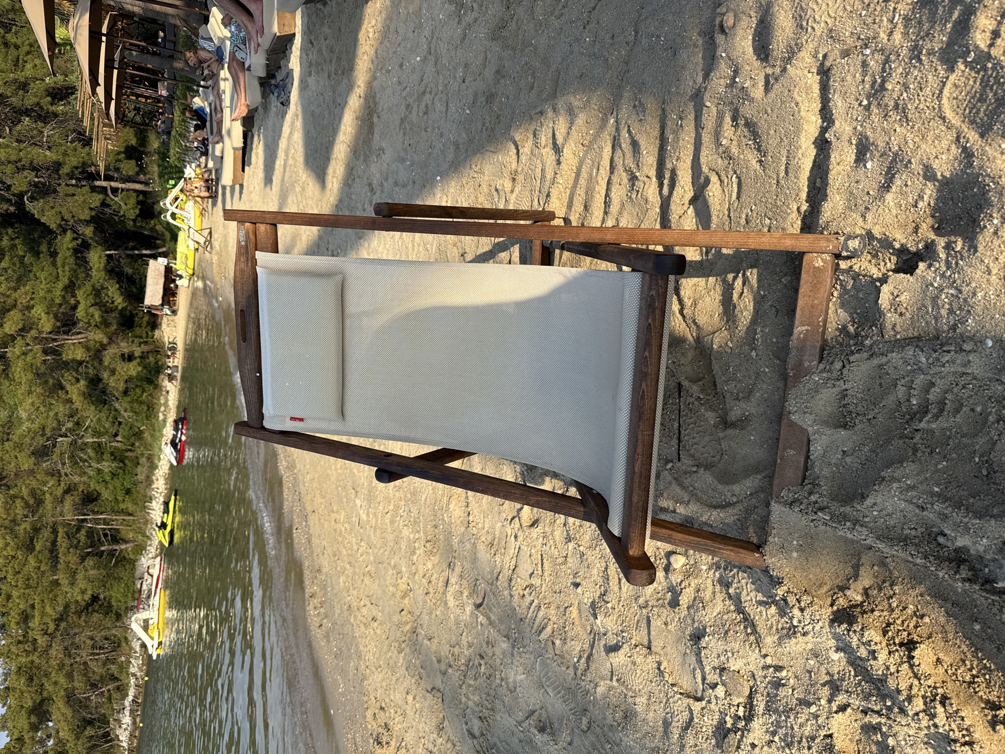 A high-angle, full shot captures a vacant, light-colored canvas beach chair made of wood, positioned on a sandy beach in front of a tranquil body of water. The beach chair is angled diagonally, with its back facing the viewer and its legs slightly dug into the sand. The beach is strewn with footprints and small pebbles, illuminated by the warm, golden light of the sun, suggesting it's either early morning or late afternoon.
In the background, the water is a calm, greenish-blue, with a few small boats and jet skis visible. Further back, a lush green forest lines the coastline, with a small, rustic hut nestled amongst the trees. To the right of the beach chair, several empty lounge chairs and umbrellas suggest a resort or beach club. The overall scene evokes a sense of peaceful relaxation.
The image depicts a scene at a beach resort, likely in the city of Thassos, Greece, given the Mediterranean flora and coastal features. The presence of jet skis and lounge chairs with umbrellas indicates a tourist-oriented location. The lighting suggests a warm, sunny day, ideal for beach activities. There are no people in the immediate foreground or engaging in any visible actions. The environment is clean and well-maintained, typical of a popular tourist destination.