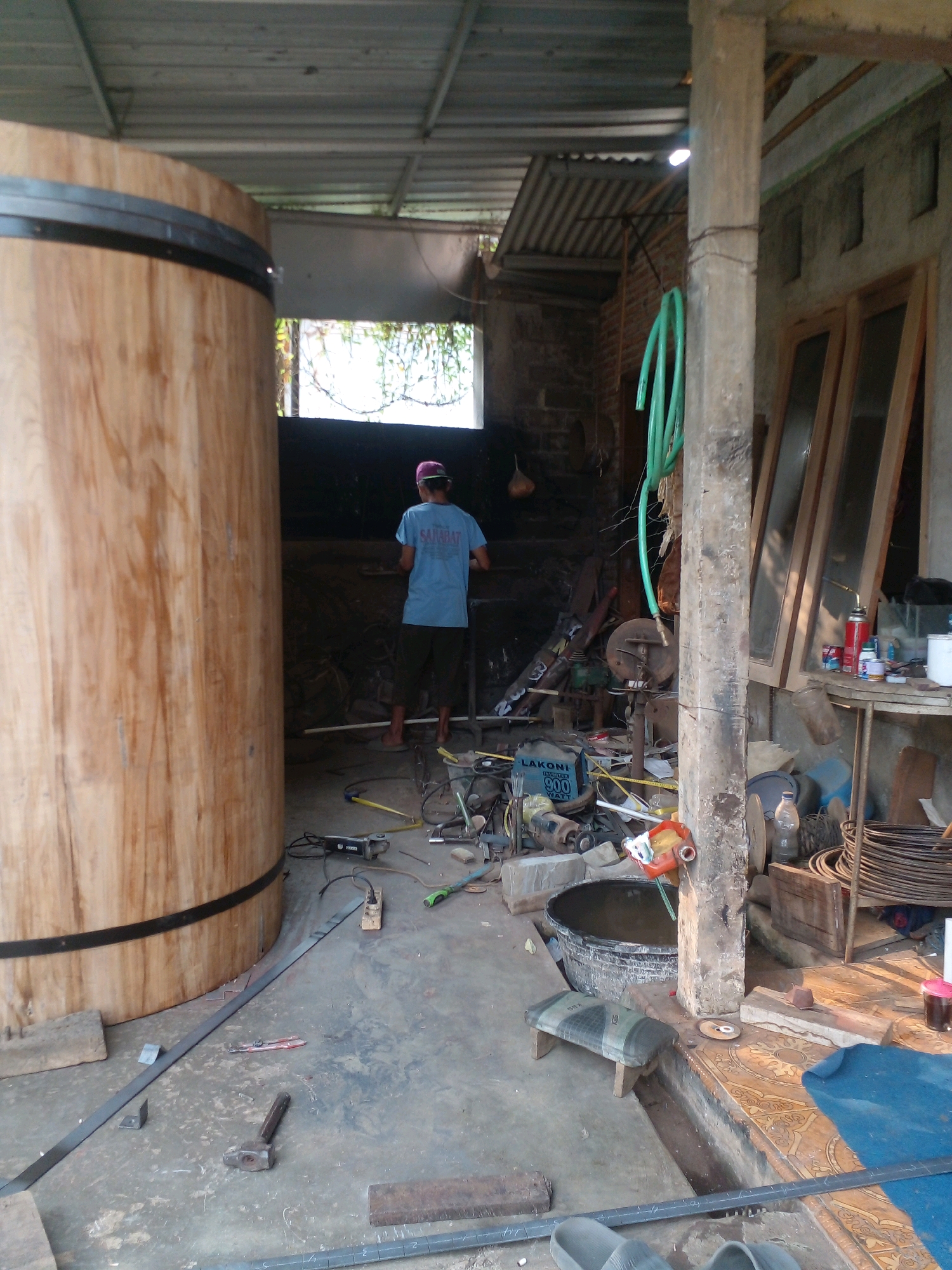 The image shows an outdoor workshop in Pungangan, Indonesia. A large wooden barrel, banded with metal, dominates the left side of the frame. A person wearing a blue t-shirt and dark shorts stands with their back to the camera, facing a cluttered workbench area in the background. The workshop is filled with tools and materials, including an angle grinder, pipes, a metal bucket, and various other items. Sunlight filters through the open roof structure, illuminating the dusty concrete floor. The overall impression is one of an active but somewhat disorganized workspace.