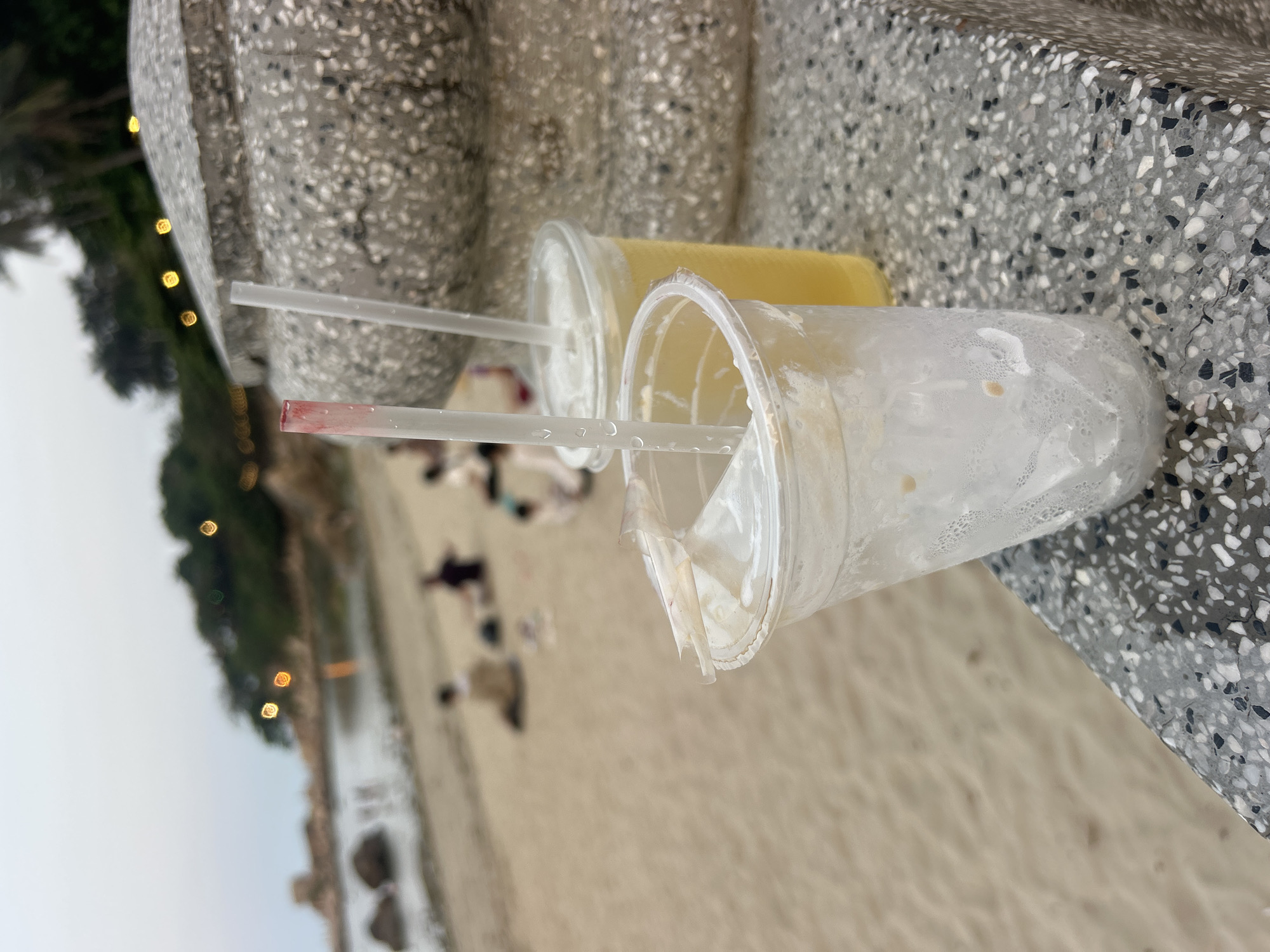 The image, taken in Bắc Nha Trang, Vietnam, presents two plastic beverage cups with straws, positioned on a speckled grey surface, likely a concrete bench or wall. One cup is nearly full with a yellow liquid, suggesting a juice or iced tea, while the other is mostly empty, exhibiting condensation and residual liquid on its interior. The straw in the empty cup shows a distinct reddish stain at its tip, possibly from a fruit drink or lipstick.
In the background, a sandy beach stretches out, populated by several blurred figures of people. Beyond the beach, a body of water is visible, flanked by lush green trees. A series of out-of-focus warm lights, likely artificial, twinkle among the foliage, suggesting the photograph was taken during late afternoon, dusk, or early evening. The sky appears overcast or hazy. No text is discernible on any objects. The scene conveys a relaxed, leisurely atmosphere by the coast.
