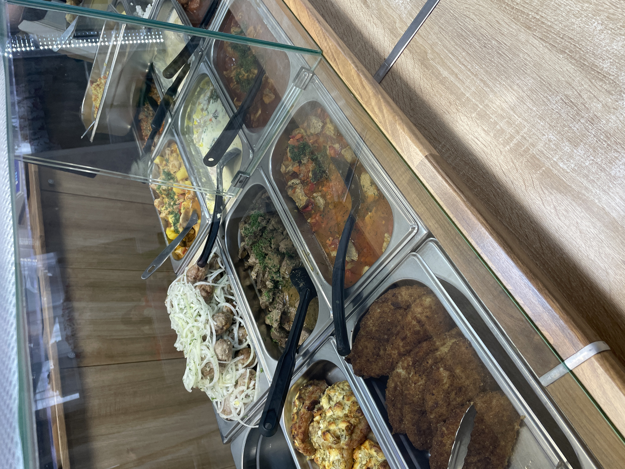 The image displays a well-stocked food service counter, likely in a cafeteria, buffet, or casual restaurant setting in Krasnoyarsk, Russia. Various cooked dishes are presented in multiple stainless steel rectangular serving trays, positioned beneath a clear glass sneeze guard. The counter surface and background feature a light-colored wooden finish.
From left to right, some of the visible food items include: a tray of small, round, possibly baked or fried items, heavily garnished with thinly sliced white onions; next to it, a darker stew containing meat chunks, possibly liver or beef, topped with green herbs. Adjacent to this is a reddish-orange stew or goulash with meat and vegetables, also sprinkled with green herbs. In the lower right foreground, a large tray holds several golden-brown, breaded cutlets. Below these, a smaller tray contains numerous irregular-shaped patties, appearing to be made of minced ingredients mixed with cheese and herbs. Black serving tongs are present in most of the trays for self-service or staff use.
The scene suggests an indoor environment, illuminated by artificial lighting, with reflections visible on the glass barrier. No individuals are present in the frame, and there is no visible text. The selection of hearty, meat-based dishes is typical of casual dining establishments.