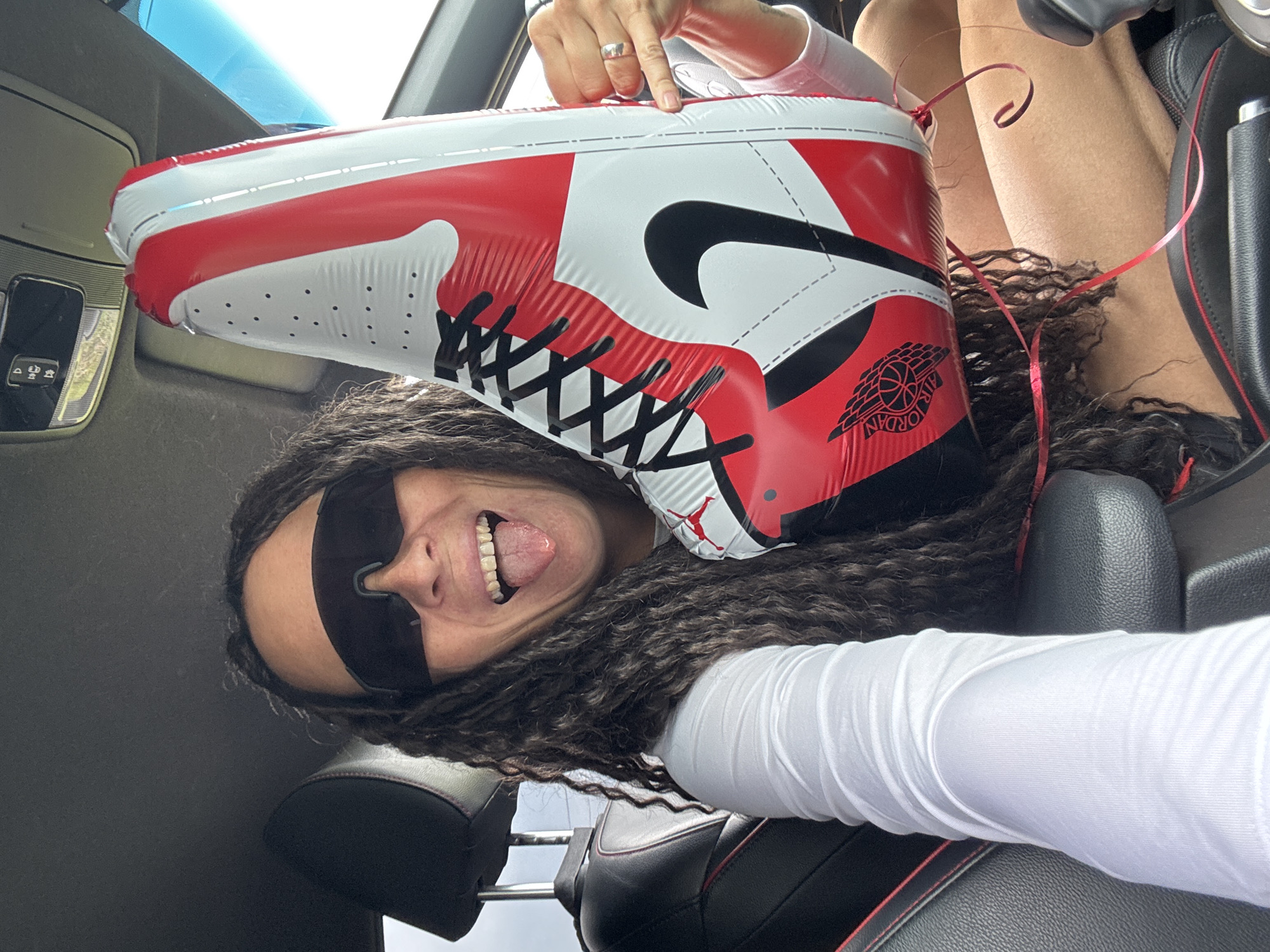 The image captures a playful scene inside a car in Chaikovsky, Russia, featuring a person with a large inflatable sneaker. The main subject is a person, likely female, with long, dark, curly hair. She is wearing black sunglasses, a white long-sleeved top, and has a ring on her finger. Her head is positioned upside down relative to the camera's orientation, with her tongue playfully sticking out and a wide smile visible. Her legs are also seen in the upper right background, confirming the inverted pose.
She is holding or posing with a prominent inflatable replica of a high-top sneaker, specifically an Air Jordan 1. The inflatable shoe is colored in classic red, white, and black. Key details like the black shoelaces, the black Nike "Swoosh" logo on the side, and the "Jordan Wings" logo on the ankle flap (which includes the text "AIR JORDAN") are clearly depicted. Red ribbons are attached to the inflatable shoe, adding to its celebratory feel.
The setting is the interior of a vehicle, evidenced by the black leather car seats with distinctive red stitching, visible both near the person and in the background. Parts of the car's ceiling and an overhead console are also seen. The bright lighting suggests it is daytime. The overall impression is one of fun and humor, with the person's expression and the unusual perspective contributing to the lighthearted mood.