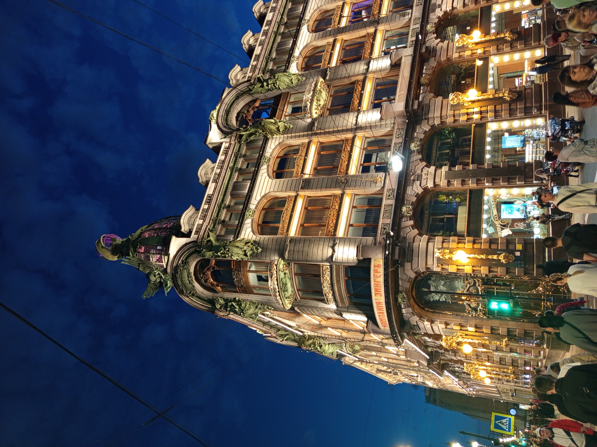 An eye-level, low-angle, nighttime shot captures the Singer Building in Saint Petersburg, Russia, from a street-level perspective. The ornate Art Nouveau architecture of the building dominates the frame, with its illuminated windows, intricate detailing, and a prominent glass dome topped with an eagle statue. The dark blue sky with scattered clouds forms the backdrop.
The street is bustling with pedestrians, many walking towards or away from the building. Some are engaged in conversation, while others are simply moving through the scene. A traffic light is visible and illuminated green. Street lamps cast a warm glow on the sidewalk and the building's facade. Several motorcycles are parked on the side of the street. A pedestrian crossing sign is also visible. The overall atmosphere is that of a lively urban environment at night.