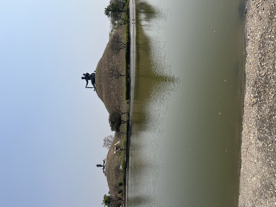 The image shows two mounds in a park, both topped with statues. One mound is on the left and the other is on the right, both are separated by a still body of water. The mounds are covered in grass and there are trees behind them. There is a stone path along the edge of the water. The image was taken from the ground level, looking towards the mounds. The location is a park in Sahibzada Ajit Singh Nagar, India. The coordinates provided locate the image within the city of Sahibzada Ajit Singh Nagar, India.