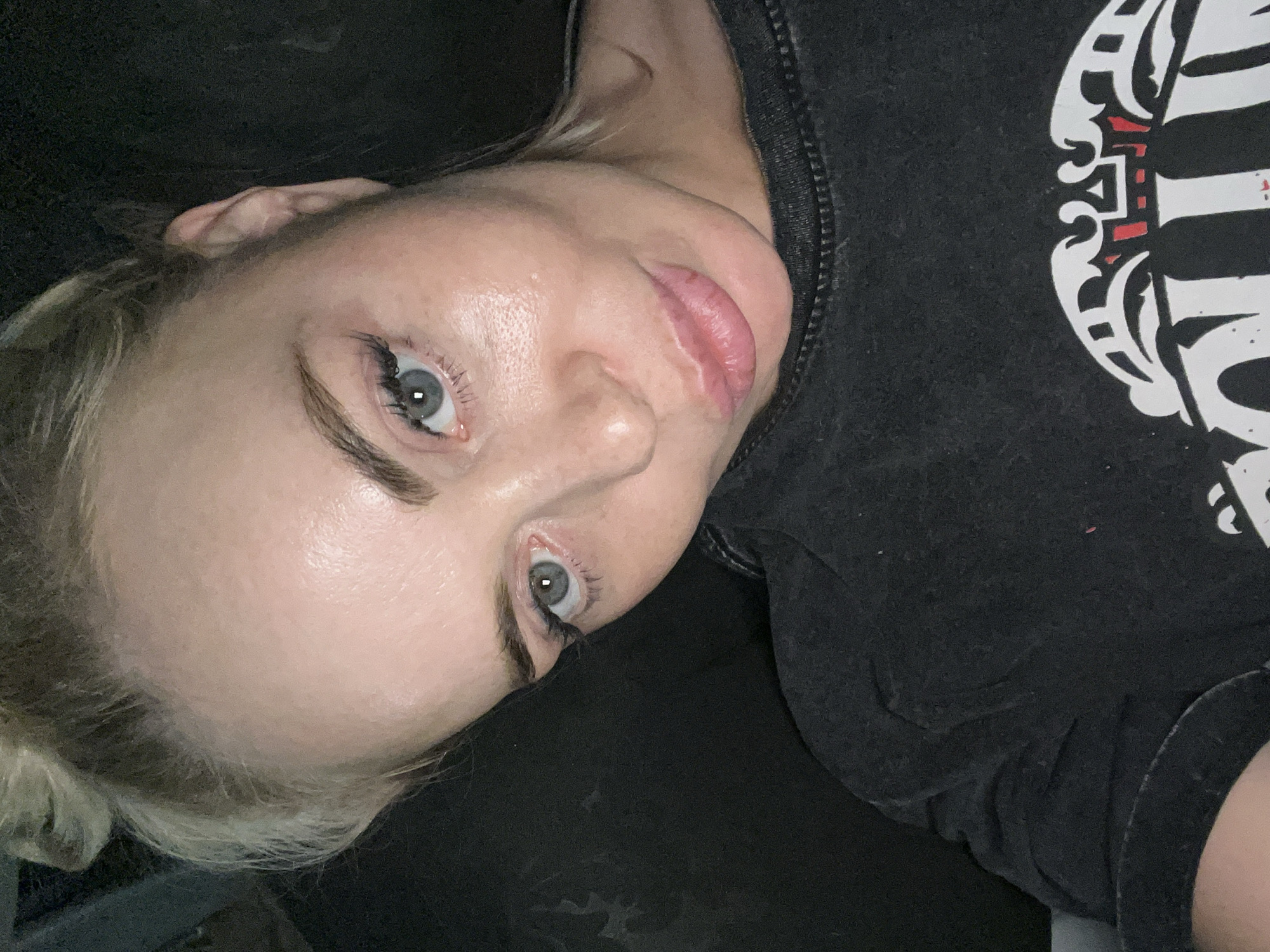 The image is a close-up, high-angle selfie of a fair-skinned woman with blonde hair styled in two buns. She is wearing a dark gray t-shirt with a white and red graphic design on it. The woman's eyes are open and looking directly at the camera, and her lips are slightly parted. Her skin appears to be shiny, possibly due to makeup or sweat. The background is dark and out of focus, suggesting an indoor setting with low lighting.