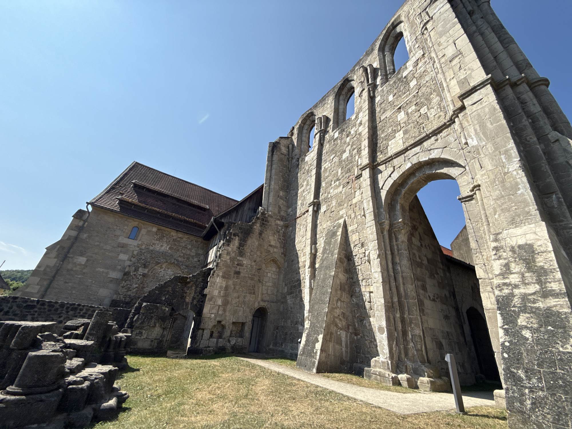 The image shows the exterior of the Walkenried Abbey ruins in Walkenried, Germany. The main subject is the weathered stone structure of the abbey, with its arches and crumbling walls visible against a clear blue sky. A portion of a modern roof with red tiles is attached to a stone building to the left. The ground is covered with dry grass and a paved path leads towards the abbey. In the foreground, there are scattered stone ruins, possibly remnants of columns or architectural elements. The lighting suggests it is a sunny day.