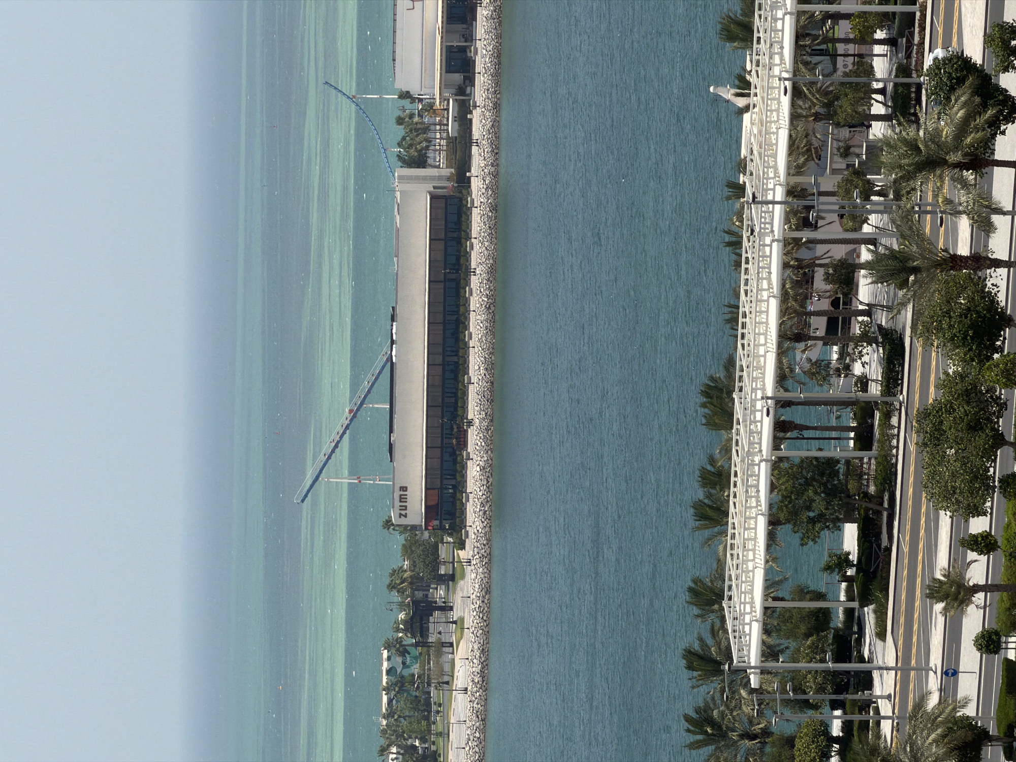 This is an eye-level, medium shot image of a waterfront area in an unknown city. In the foreground, a calm, turquoise body of water is visible. Beyond the water, a paved walkway lines the shore, bordered by a low, rocky wall. Behind the walkway are several palm trees and other greenery. A long, white, elevated structure with a patterned roof extends across the midground. Further back, a low-rise building with large windows is visible, with the word "zuma" written in dark letters above the entrance. To the right of this building is another structure, partially visible. In the background, the ocean stretches to the horizon under a hazy sky. A faint, light blue roller coaster track curves across the upper right quadrant of the image, extending from the far left. The scene appears to be a commercial or recreational area.