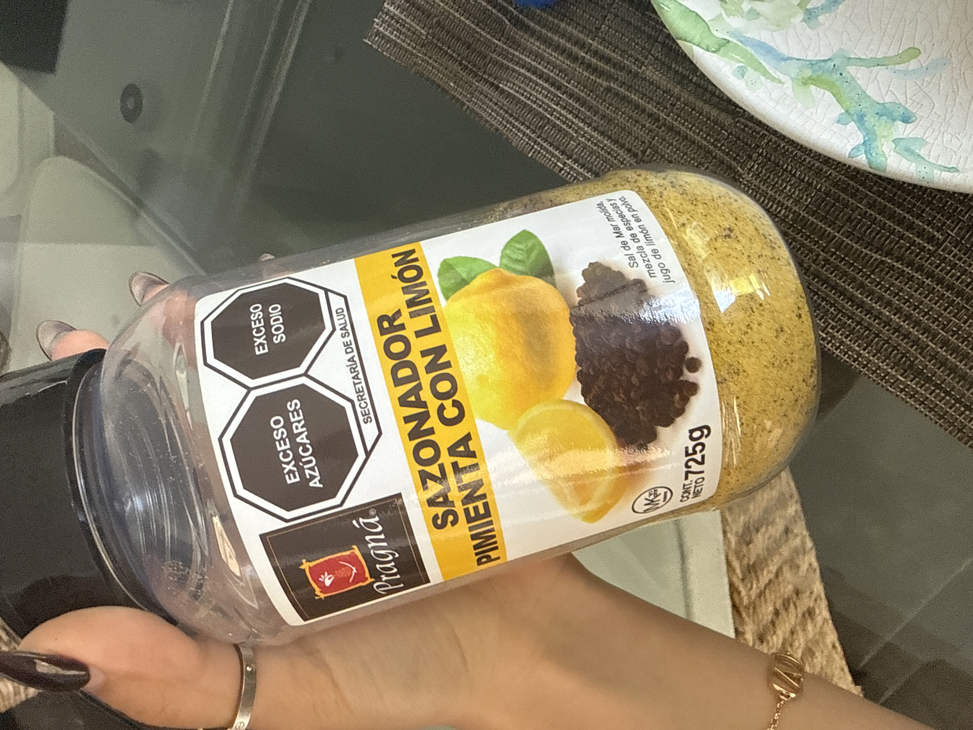 A person is holding a large, clear plastic container of "Pragná" brand seasoning in an indoor setting in San Bernardino Tlaxcalancingo, Mexico. The container, filled with a yellowish, granular spice mixture, is prominently labeled "SAZONADOR PIMIENTA CON LIMÓN" (Seasoning Pepper with Lemon).
The person's hands and forearms are visible, with long, dark-painted fingernails and two bracelets: a silver one on the left wrist and a gold-colored chain bracelet on the right. The container is angled, allowing a clear view of its label. The background features a glass table with a textured brown placemat. Part of a white ceramic plate with a light blue and green abstract design is visible on the right side, resting on the placemat.
Visible text on the container includes health warning labels: "EXCESO AZÚCARES" (Excess Sugars) and "EXCESO SODIO" (Excess Sodium), along with "SECRETARÍA DE SALUD" (Ministry of Health). The brand name "Pragná" is displayed with a red logo. Further text details the ingredients as "Sal de Mar molida, mezcla de especias y jugo de limón en polvo" (Ground Sea Salt, spice blend, and lemon juice powder). The net weight is indicated as "CONT. NETO 725g." The label also features an illustration of two lemons and black peppercorns. The scene depicts a close-up view of the product in what appears to be a domestic kitchen or dining environment.