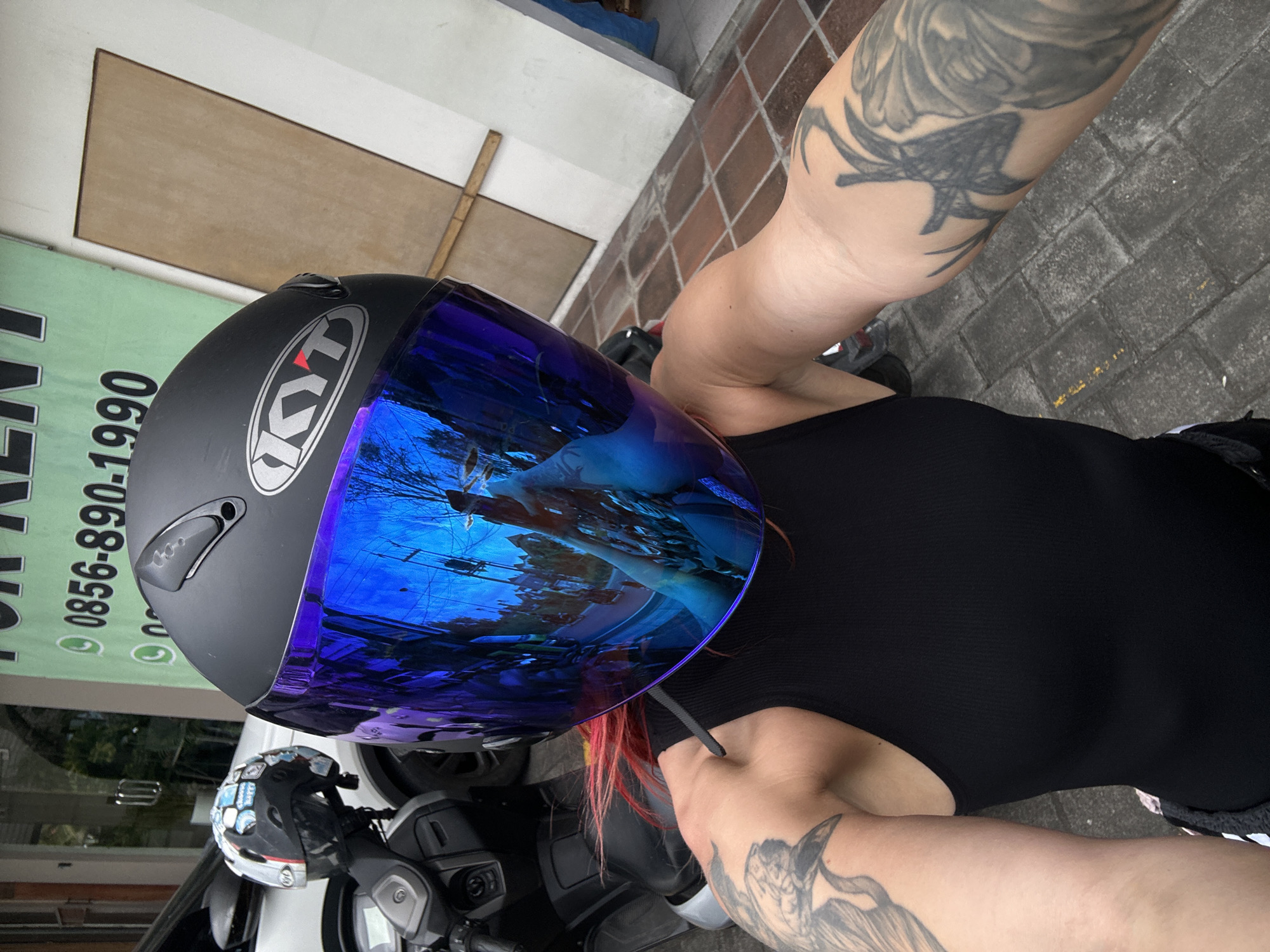 In the city of North Kuta, Indonesia, a person wearing a black sleeveless shirt and a black motorcycle helmet with a blue reflective visor is positioned in the foreground, facing the camera. The helmet has a silver "KYT" logo on its side. The person has tattoos on their left arm and visible on their right arm. Red-dyed hair is visible peeking out from under the helmet. A motorcycle is partially visible to the left of the person, and another helmet with stickers is attached to it. In the background, a green sign with white text, including a phone number, is displayed on a building. The ground is paved with interlocking gray bricks. The scene appears to be outdoors on a street or sidewalk.