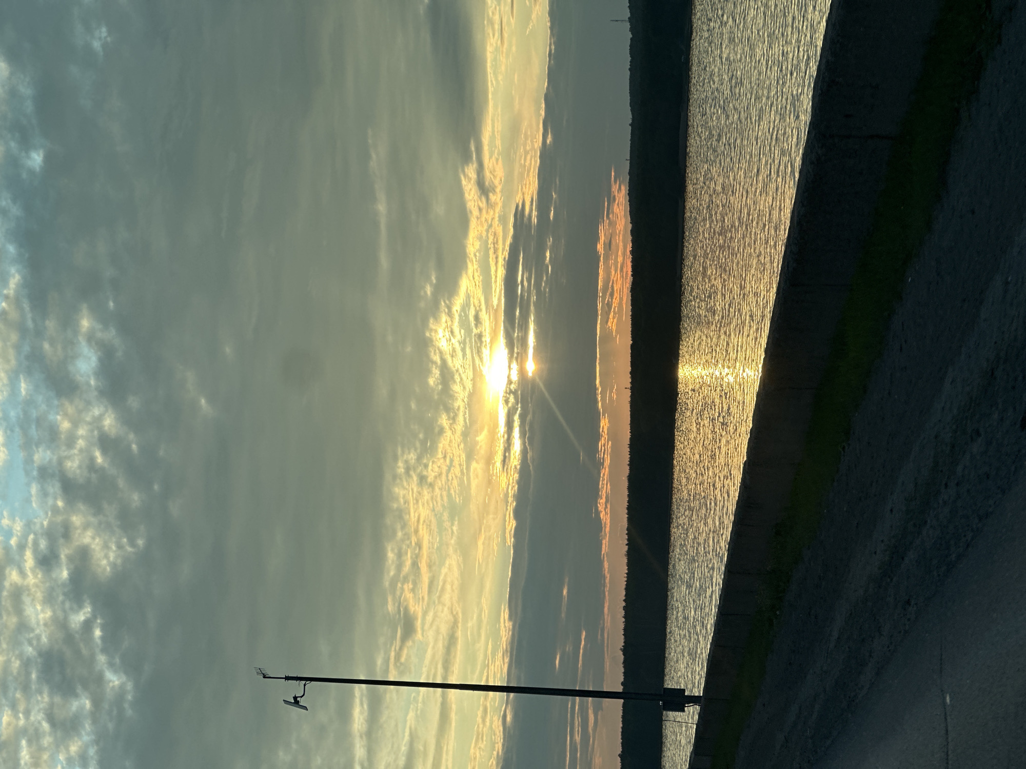 The image shows a sunset over the Kama River in Chaikovsky, Russia. The sky is filled with clouds, and the sun is setting behind them, casting a warm glow on the water. The river is calm, with gentle ripples reflecting the colors of the sky. On the left side of the image, there is a tall pole with some equipment attached to it, possibly for monitoring or communication. In the foreground, there is a dirt road leading to a concrete embankment along the river. The embankment is covered with some grass. The overall scene is serene and picturesque, capturing the beauty of nature at dusk. The location is identifiable as Chaikovsky, Russia, due to the specific geographical features and the context provided.