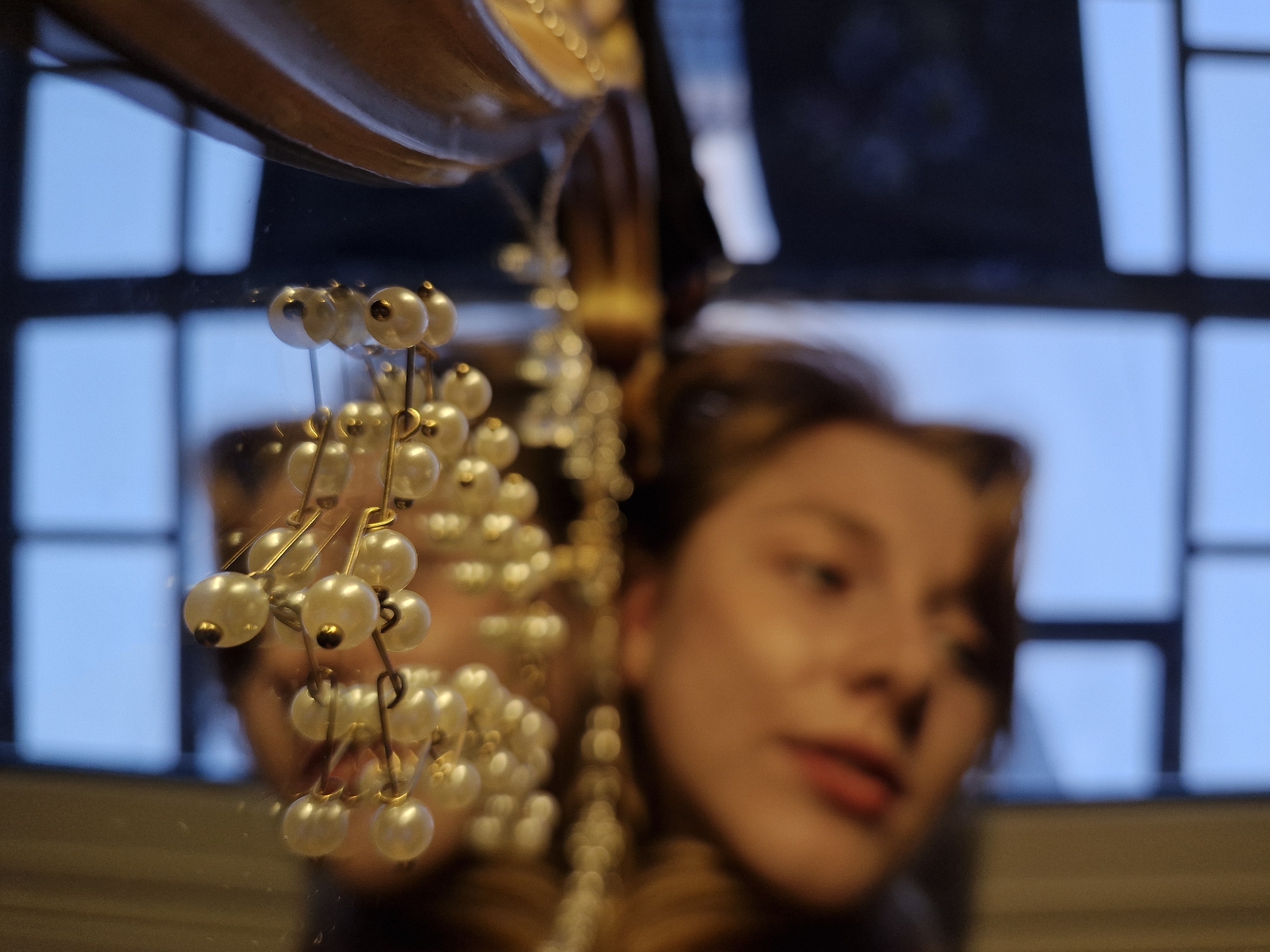 This media file presents a layered composition featuring a reflection, selective focus, and distinct objects. In the sharp foreground, a string of lustrous, pearl-like beads is prominent. These small, white spheres are mounted on golden wires, creating a delicate, dangling cluster that occupies the left and central parts of the image.
Behind and through the reflection where the beads are positioned, the softly blurred face of a young woman is visible, predominantly on the right side of the frame. Her gaze appears gentle and slightly contemplative, with a hint of a serene expression or a subtle smile. Her features are diffused, contributing to an artistic and ethereal quality.
Further in the background, also seen as a reflection, is a window characterized by a clear grid pattern, indicating multiple panes. The light filtering through the window is a cool, soft blue, suggesting either an overcast day or the quiet hours of twilight. A curved, warm-toned object, possibly made of wood or metal and resembling a piece of furniture or an architectural detail, is partially visible at the top of the image.
The image utilizes a shallow depth of field, bringing the artificial pearls into sharp focus while rendering the woman's face and the background window in a soft blur. The overall setting is an indoor environment, likely a room in Yekaterinburg, Russia. No visible text is present in the image.