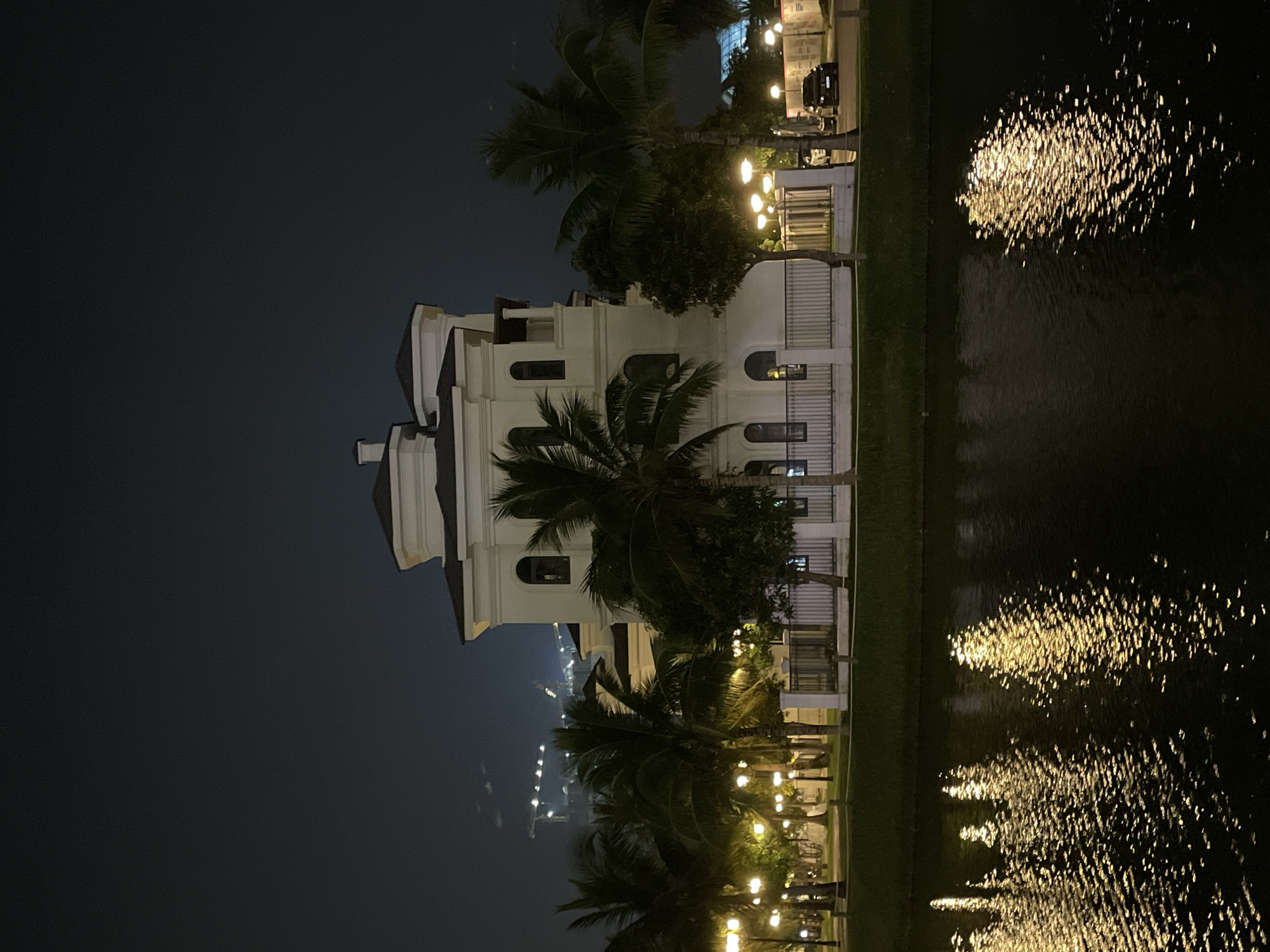 This is a nighttime photograph of a large, white, multi-story building with a distinctly modern architectural style, located near a body of water. The building has several prominent features, including a central tower with a peaked roof, arched windows, and decorative trim. Palm trees and other greenery are visible in front of the building, suggesting a tropical or subtropical climate. Several streetlights illuminate the foreground, casting reflections on the water's surface. A white fence runs along the edge of the property in front of the building, and a dark-colored car is parked on the street in the background. In the far distance, several construction cranes are visible against the dark sky, indicating ongoing development. The overall atmosphere of the image is serene and slightly dramatic due to the nighttime lighting. Based on the presence of palm trees and the architectural style, this building is likely situated in a planned community or resort area within Hà Nội, Vietnam, possibly near the Red River or in a developing suburban district.
