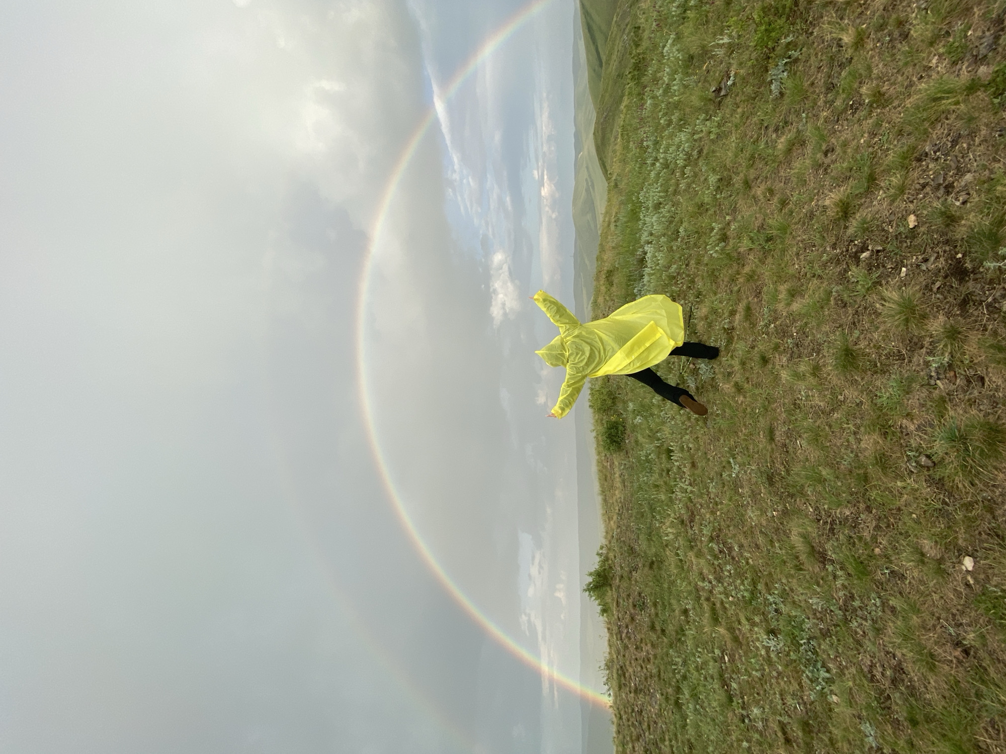 A person wearing a yellow raincoat stands with their arms outstretched on a grassy hill, facing a double rainbow in a cloudy sky. The person's back is to the camera. The landscape features rolling green hills and a body of water in the distance. The image appears to be taken in an outdoor, natural environment.
