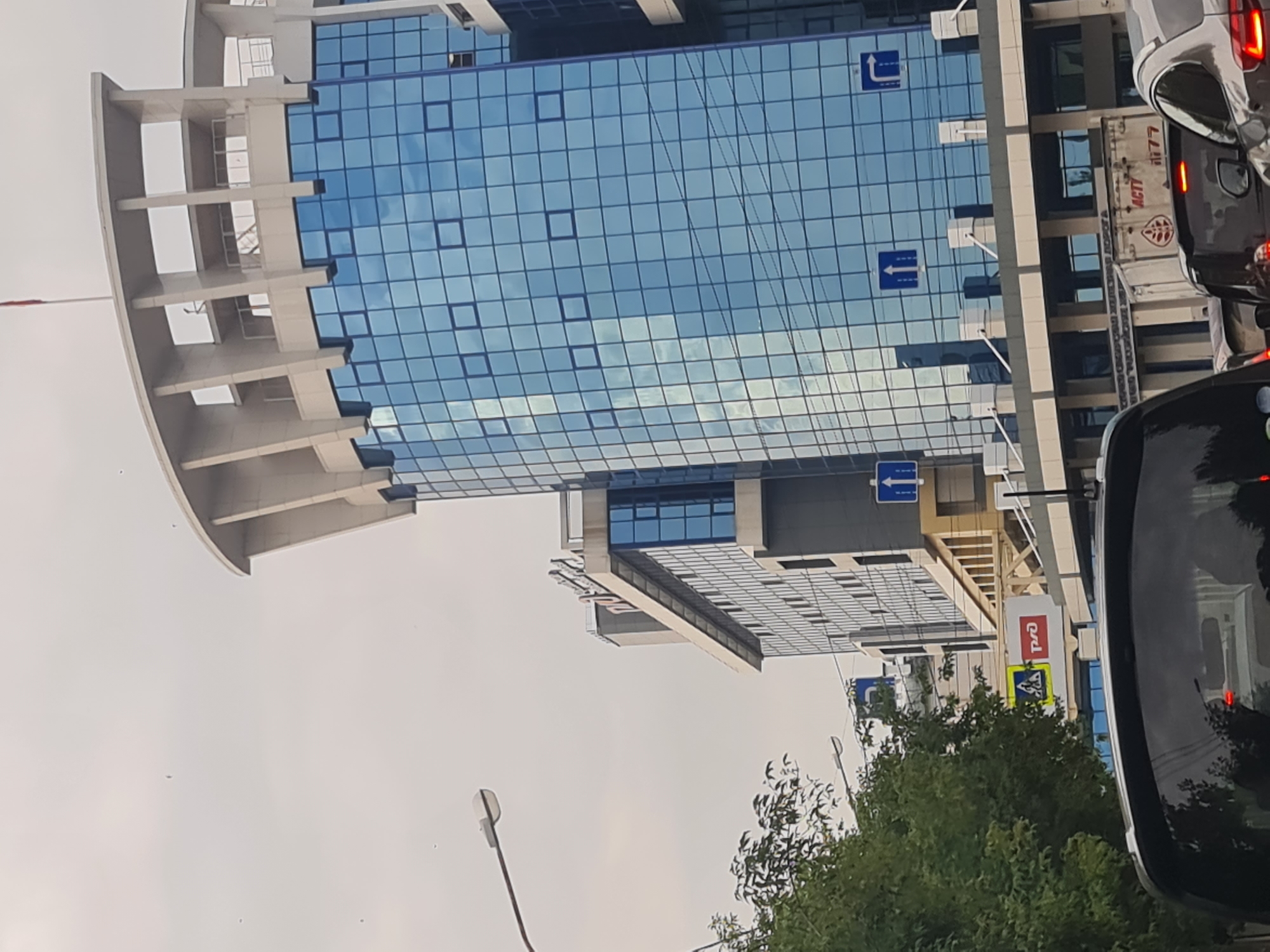 The image shows a street view in Irkutsk, Russia, dominated by modern architecture. In the foreground, the rear of a dark-colored car is visible, suggesting the photo was taken from a vehicle in traffic. To the right, another car's reflection can be seen on the car in front.
The mid-ground is filled with buildings. A tall, prominent building with a curved top features a facade of reflective blue glass windows, arranged in a grid pattern. The building's structure includes white concrete pillars and upper levels. Adjacent to it, another building, appearing older or of a different design, has a dark gray exterior with numerous rectangular windows. A sign with Russian text and a red rectangle is visible on this building.
Traffic signs are present, including a blue square sign with a white upward-pointing arrow and a blue square sign with a white arrow indicating a right turn. A pedestrian crossing sign with a yellow triangle and a yellow diamond is also visible.
A lamppost with a modern, minimalist design extends from the left side of the frame. Trees with green foliage are situated on the left, partially obscuring the view of the buildings. The sky is overcast and a pale gray. A red flag flies from the top of the main building. A truck with Russian text and branding, including "ACTY" and the number "77", is visible behind the cars. The overall scene depicts an urban environment, likely a commercial or business district, under a cloudy sky.