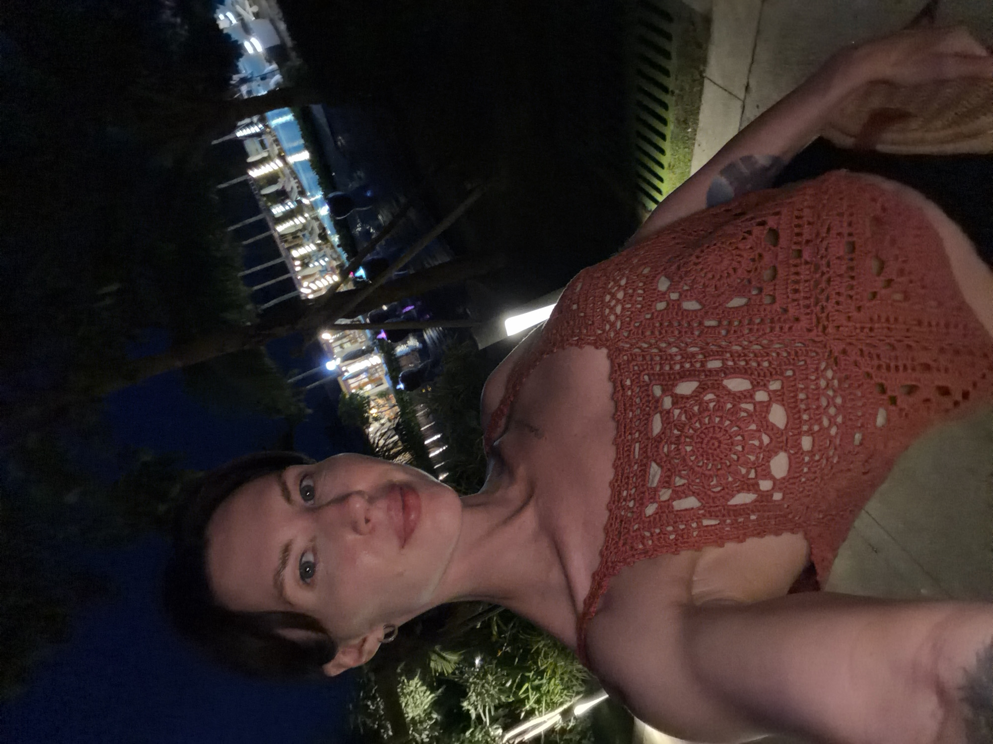 A woman with short brown hair and light skin poses for a selfie at night. She is wearing a red crocheted tank top. Her left arm is bent, resting on her lap. The background shows a dimly lit outdoor area with palm trees and a swimming pool illuminated by lights. The overall setting appears to be a resort or hotel. The location is identified as Dong Ninh Hoa Ward, Vietnam.