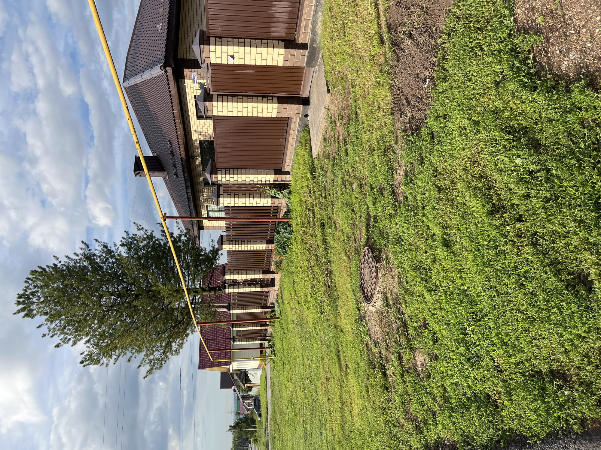 The image depicts a residential property with a brick and brown metal fence. A mature pine tree stands to the left of a house with a dark red roof. A yellow gas pipe runs horizontally across the frame, supported by metal posts. In the foreground, a patch of green grass covers the ground, with a circular metal manhole cover visible. In the background, other houses and a road can be seen, along with power lines and a cloudy sky. The location is Ufa, Russia.