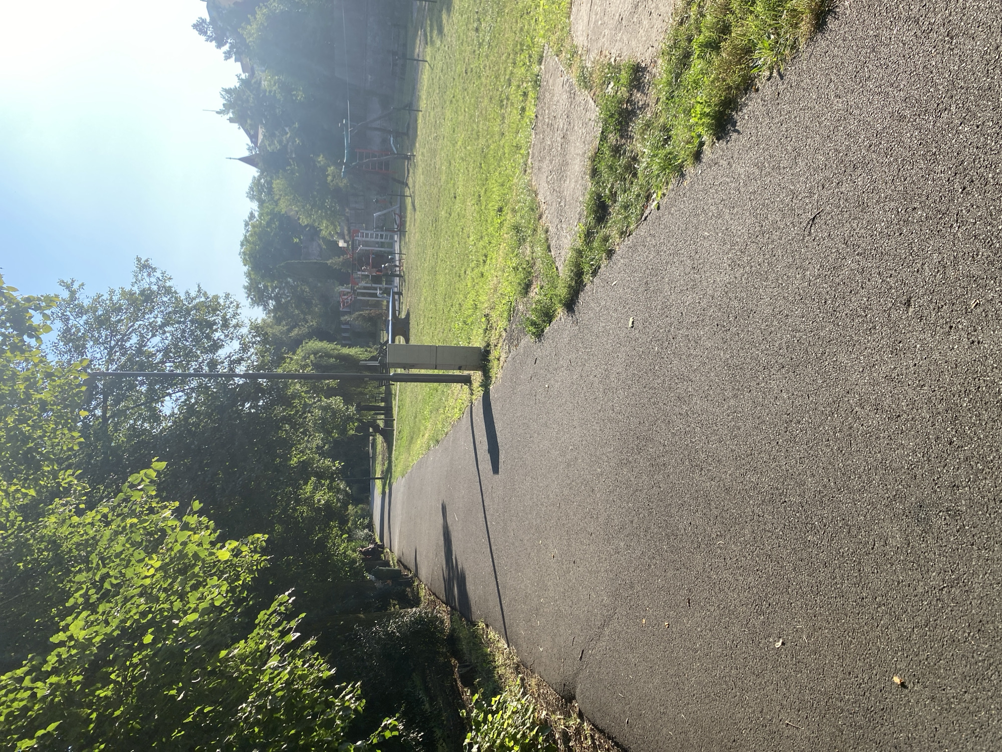 An asphalt pathway curves through a park-like setting under a bright, clear sky. Lush green trees line the left side of the pathway, their leaves catching the sunlight. To the right, a grassy area features a children's playground with swings and climbing structures. In the background, buildings with distinctive architecture, possibly historical, are visible amidst more trees. A lamppost stands near the pathway, and a concrete utility box is positioned beside it. A person is seated on a bench in the distance on the left. The scene suggests a well-maintained urban park.