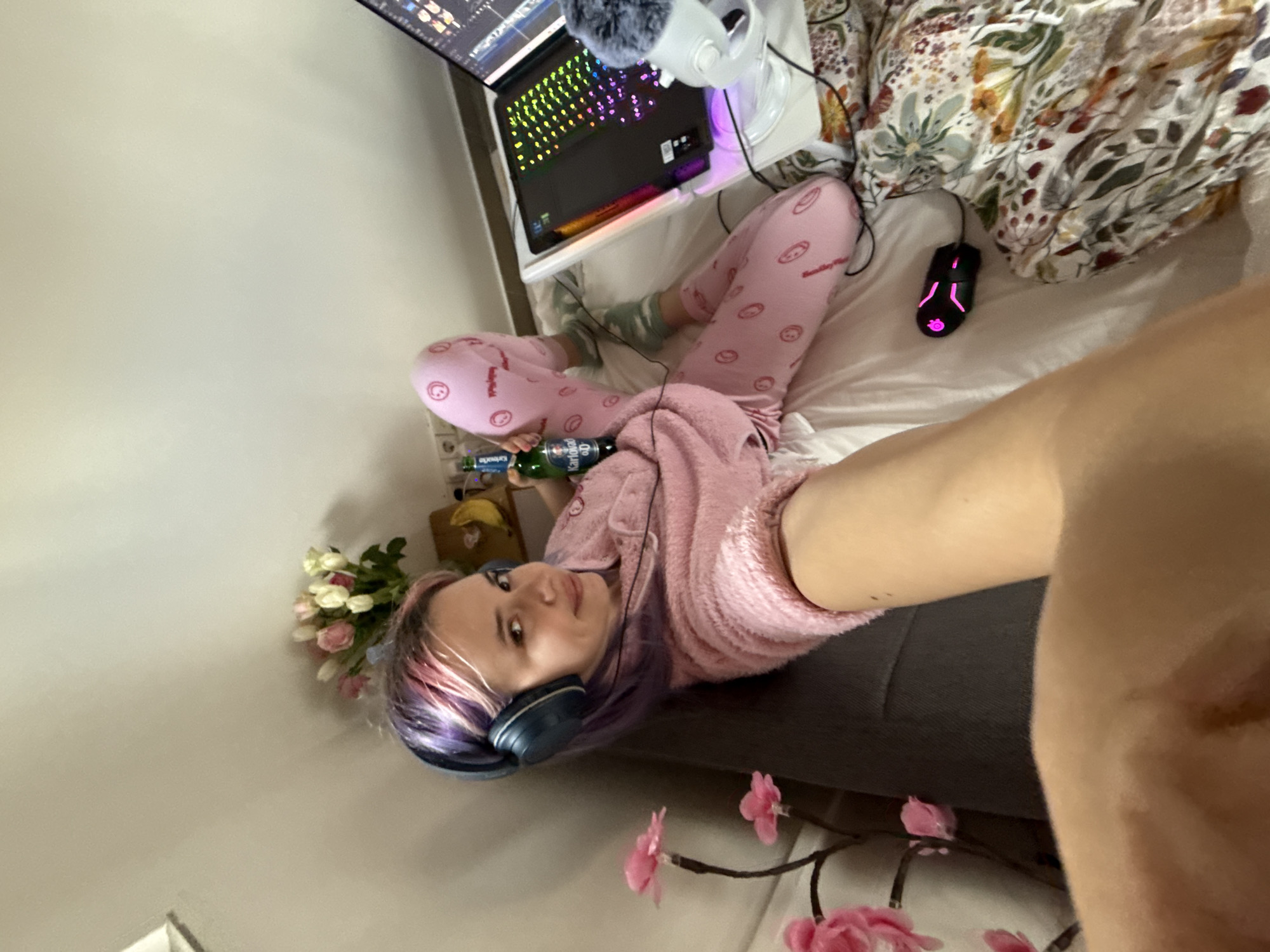 An overhead, slightly angled view shows a female individual in a bedroom setting, likely in the City of Zagreb, Croatia. The subject has multi-colored hair (purple and pink) and is wearing dark blue over-ear headphones, a pink fuzzy long-sleeved top, and pink pajama pants with circular patterns. She is lying on her back, propped up on a bed with white sheets and a floral duvet, her legs bent. She holds a green glass bottle labeled "Karlovačko 0.0" in her right hand and looks directly at the camera with a slight smile. To her right, a black laptop with a multicolored RGB keyboard is open on a white bed tray, displaying what appears to be video editing software. A white USB microphone with a grey fuzzy windscreen is positioned near the laptop. A black gaming mouse with pink LED accents rests on the bed. In the background, behind the subject, a bouquet of pink and white tulips is visible on a surface, next to a yellow banana. A decorative lamp with pink floral elements stands on the left side of the frame. The walls are light-colored.