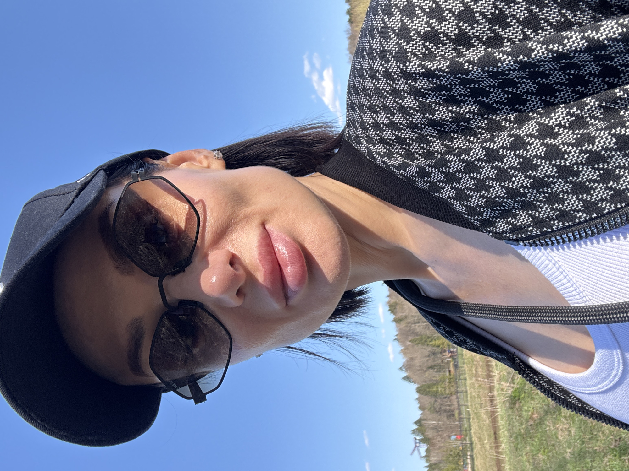 A woman wearing sunglasses and a cap is angled upward against a bright blue sky in an outdoor setting. She is wearing a black and white patterned jacket over a white top. The background includes a field with sparse trees and grass. The location is Unknown, Unknown.
