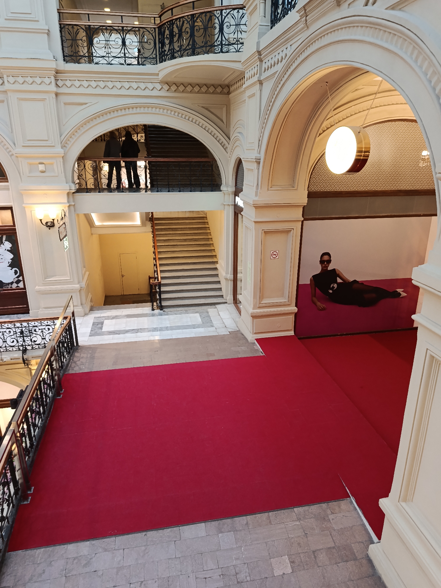 The image depicts an interior view of GUM (State Department Store) in Moscow, Russia. The architecture is ornate, featuring arched passageways, decorative railings, and classical details. The scene is viewed from an elevated position looking down. A red carpet extends across the foreground and into the midground.
A large advertisement featuring a reclining woman in a black dress and sunglasses is visible under an archway. Two people are standing on a balcony on the left side of the image, above a staircase. A circular light fixture hangs from the archway above the advertisement. There are ornate railings on the left side of the image and two levels of balconies on the left. A "no smoking" sign is next to the advertisement.