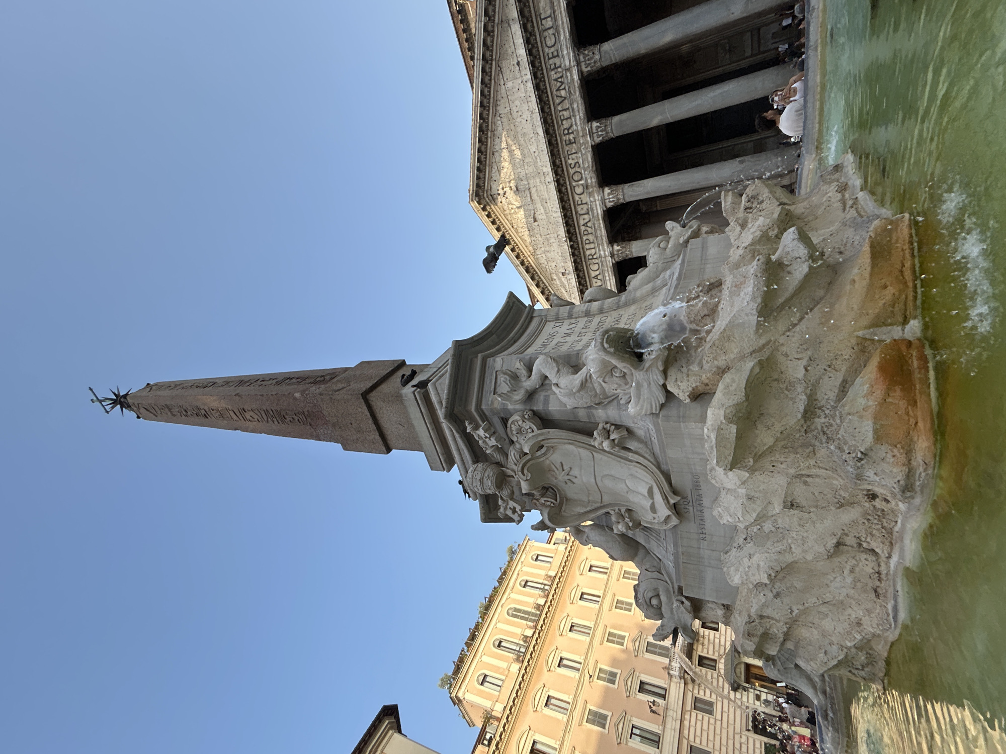This low-angle, portrait-oriented photo captures a section of the Fountain of Neptune in Piazza Navona, Rome, Italy. The central subject is the impressive fountain's sculptural elements, including an obelisk topped with a star-like finial and ornate carvings of mythological figures and coats of arms. Water is actively flowing from the fountain, creating a dynamic and refreshing visual.
In the background, the majestic facade of the Pantheon, with its massive columns and pediment, is visible against a clear blue sky, suggesting fair weather. The Pantheon's inscription, "LAGRIPPA LF-COSTERTIVM FECIT," is partially legible. Below the fountain, a charming cream-colored building with numerous windows lines the square, hinting at the urban setting.
A few people are scattered around the fountain and in front of the Pantheon, appearing as small figures engaged in sightseeing or enjoying the public space. The time of day appears to be late afternoon or early evening, as indicated by the warm lighting on the buildings and the clear, bright sky. The overall atmosphere is one of historical grandeur and lively public activity, characteristic of a popular Roman piazza. Text visible on the fountain reads "RESTAURATA 1880."