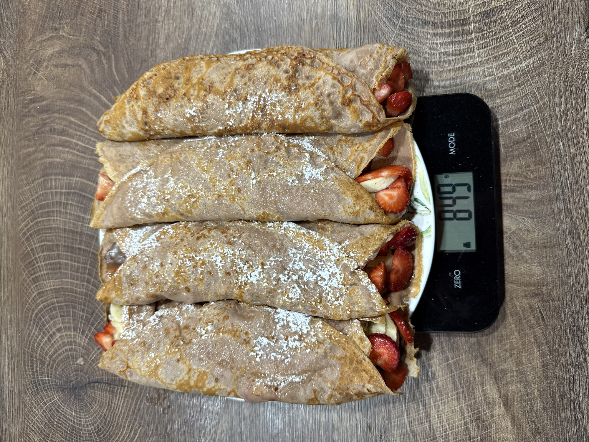 The image originates from an indoor setting in the city of Sofia, Bulgaria. It captures an overhead view of five rolled crepes, lightly dusted with powdered sugar, arranged on a white plate with a subtle floral or leaf pattern on its rim. Each crepe is filled with sliced strawberries and banana pieces, visible at their open ends. The plate rests on a black digital kitchen scale, which displays "849 g" on its screen. The scale also features "ZERO" and "MODE" buttons. The entire arrangement is situated on a wooden surface with a distinct, light brown grain pattern.
