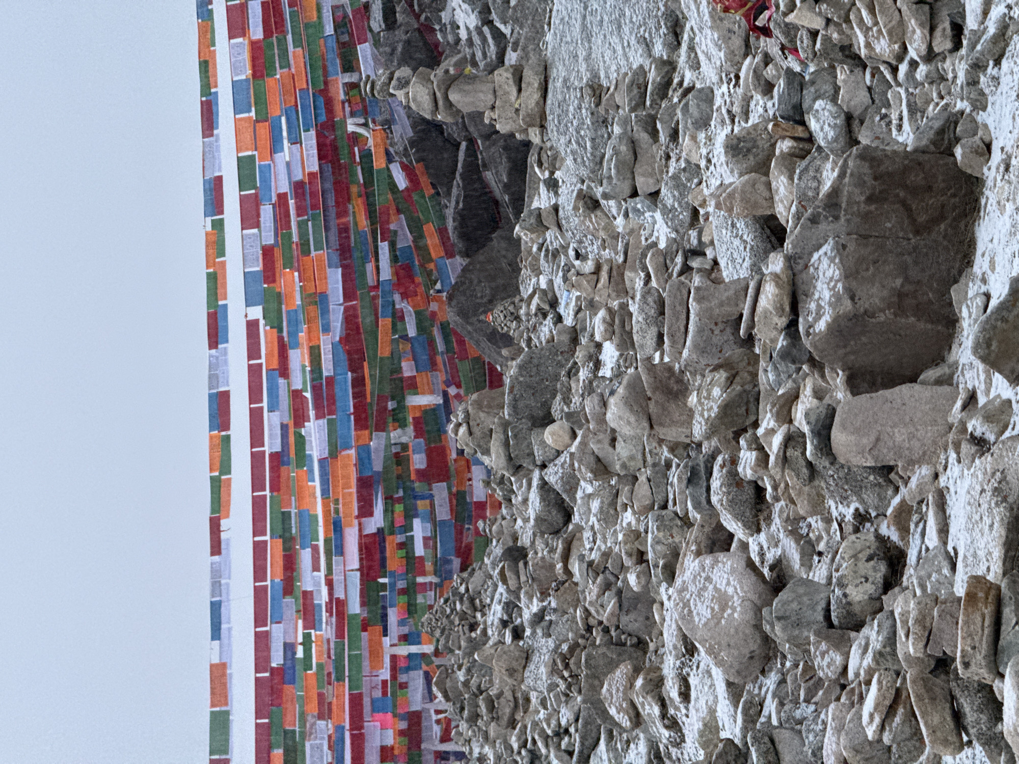 The image depicts a serene and culturally significant outdoor scene, likely a mountain pass or sacred site. On the left side of the frame, numerous multi-colored prayer flags, known as lungta in Tibetan Buddhism, hang vertically. These rectangular flags are strung in rows and feature a repeating pattern of colors including red, orange, blue, green, and white. They appear somewhat weathered, indicating exposure to the elements.
The right side of the image is dominated by a rugged, grey rock face or slope, composed of countless individual stones and larger boulders. Among these natural formations, several cairns, or stacks of stones, are visible. These carefully balanced stone piles suggest human interaction with the environment, often as a form of offering, prayer, or trail marking in such regions. The overall texture of the rocks varies, with some areas appearing smooth and others rough or fragmented.
The sky, visible on the far left behind the prayer flags, is a light, overcast white or very pale blue, indicating daylight, possibly a cloudy day or bright morning/afternoon. No specific time of day can be definitively determined, but the lighting is even. There is no visible activity involving people, but the presence of the flags and cairns points to spiritual or cultural practices. No discernible text is visible on the flags or stones.
The visual cues, particularly the prayer flags and stone cairns, strongly suggest a location in the Himalayas or a region with Tibetan cultural influence. While the provided context states "Parga town, China," this description does not align with the typical imagery associated with Parga (a town in Greece) and instead points towards areas like Tibet or regions within China that share Tibetan cultural practices.