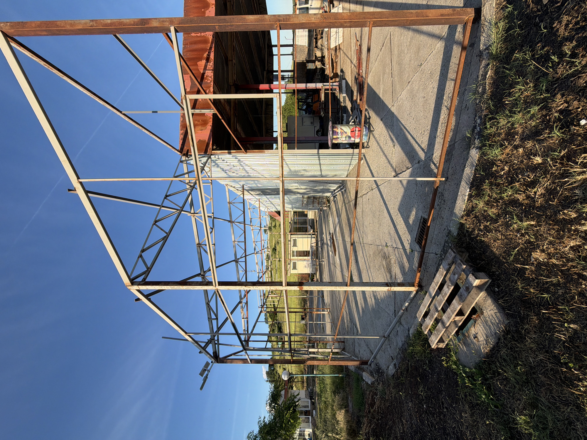 The image depicts a partially constructed metal frame structure, possibly for a greenhouse, located outdoors in Dabnik, Bulgaria. The structure is predominantly composed of rusted metal beams connected to form a series of triangular and rectangular supports. The frame stands on a concrete slab or foundation adjacent to an area with grass and vegetation. In the background, there is a building with corrugated metal siding, as well as other structures and greenery. A wooden pallet rests near one corner of the frame. The sky is visible through the frame, suggesting an open construction. The location is open and appears to be an industrial or semi-industrial area on the outskirts of Dabnik. No people are visible in the image.