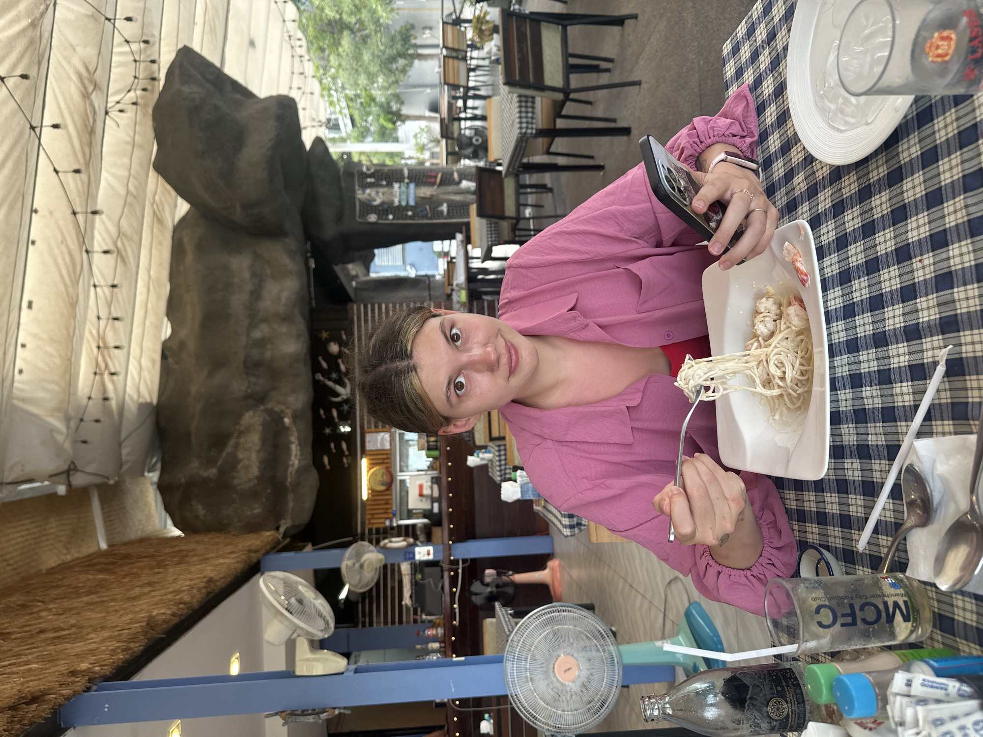 A woman is photographed at a table laden with food and utensils, seated outside a restaurant in Baan Chaweng, Thailand. She holds a smartphone in her left hand and a fork with noodles in her right. The table is covered with a blue and white checkered tablecloth. A plate of shrimp pasta is to her right, and a plate and a glass of water sit at the upper right of the table. More bottles, cutlery and paper napkins are nearby. Three fans are visible in the foreground, while behind her, other tables and chairs are arranged outdoors. A rock structure and a roof are also partially visible.