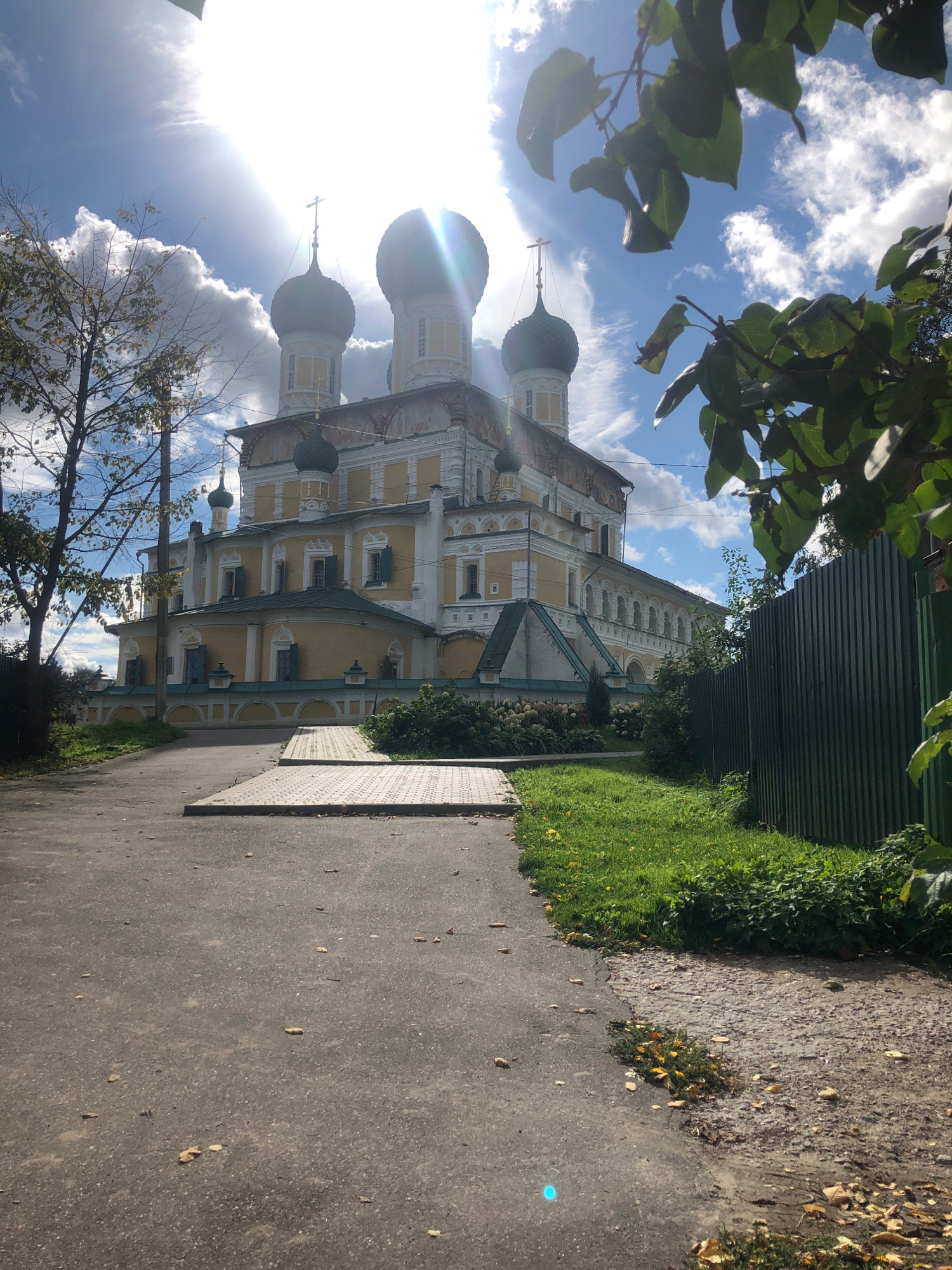 A view of the Resurrection Cathedral in Tutaev, Russia, shows its yellow facade, white architectural details, and five onion domes against a partly cloudy blue sky. Sunlight streams from the upper left, creating lens flare and illuminating the clouds. The cathedral is framed on the right by a tall, dark green corrugated metal fence and foliage from a tree with green leaves in the foreground. To the left of the cathedral, a bare tree with sparse autumn leaves stands near a utility pole. A paved path leads toward the cathedral, with fallen leaves scattered on it and the adjacent grass. A portion of the path consists of metal plates. The ground slopes down from the foreground toward the cathedral.