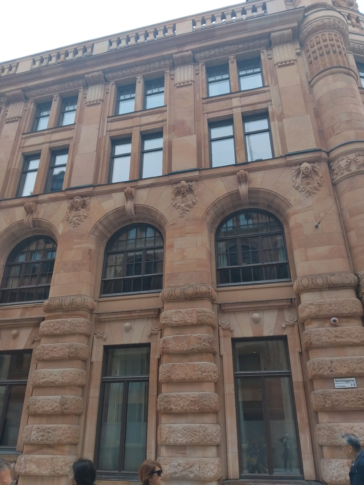 This is a low-angle, eye-level shot of the exterior of a grand, historic building made of sandstone. The building has multiple stories, with numerous windows of various sizes and shapes. The architectural style is ornate, featuring decorative carvings around the windows and along the roofline. There are prominent stone columns on the lower floors with decorative patterns. Some of the windows are arched, and others are rectangular. A street sign is visible on the right side of the building, reading "Drottninggatan 1-5" and a smaller text "Ly. Rosenblad". This indicates the location is Drottninggatan in Stockholm, Sweden. There are at least three people visible in the lower part of the frame, appearing to be pedestrians on the street. One person is facing towards the building, another is in profile facing right, and a third person is mostly obscured. The overall impression is one of an impressive, well-preserved urban architectural landmark.