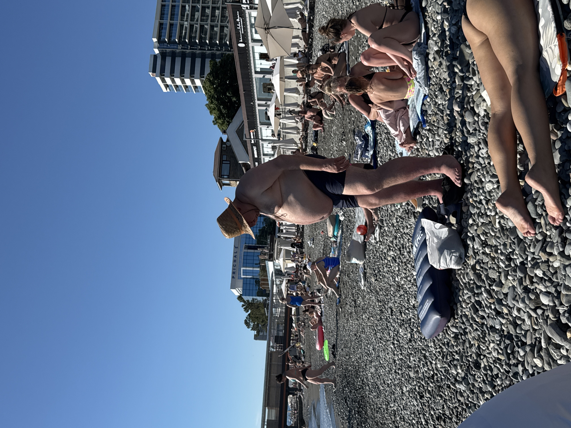 The image depicts a crowded beach in Sochi, Russia, on a clear, sunny day. The beach is covered in pebbles, and several people are sunbathing, walking, or relaxing on towels and inflatable mats. In the foreground, a man wearing a straw hat and swim trunks is visible from the chest down. He appears to be standing and facing away from the camera. In the background, there are several buildings, including a multi-story hotel and a building with the word "PRICHAL" on it, suggesting a pier or waterfront location. Beach umbrellas and loungers are scattered across the beach, indicating a popular tourist area. The sea is visible in the distance, with gentle waves lapping at the shore. The overall scene is one of a typical summer day at a beach resort.