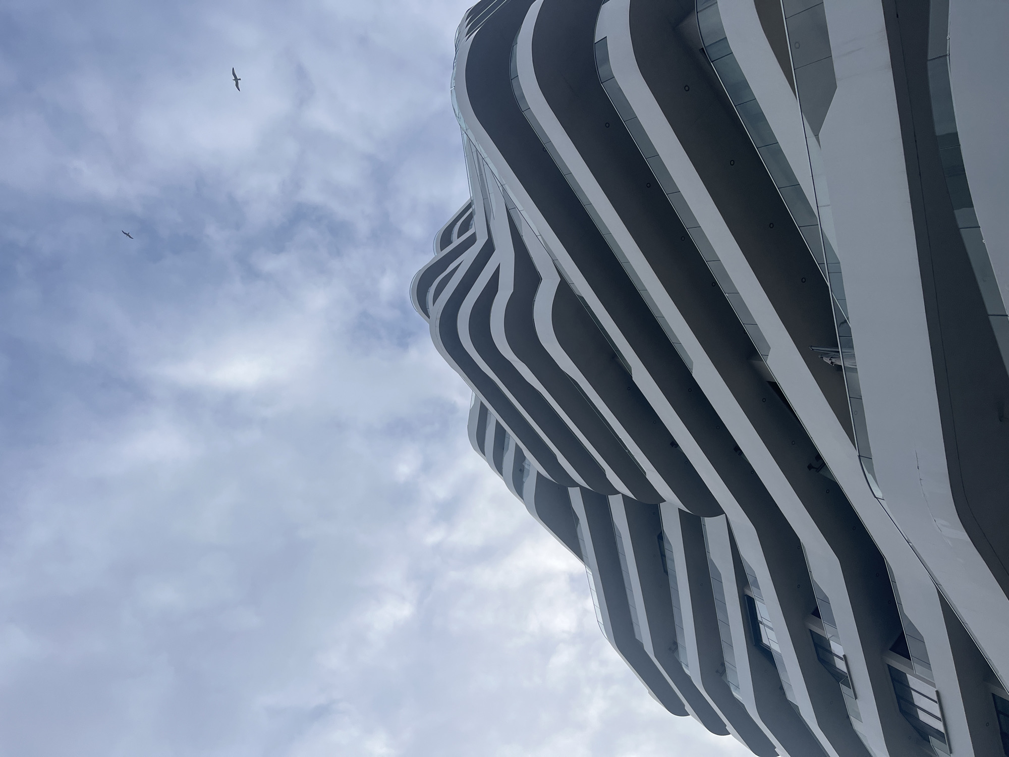 The image shows a view from below of a modern building with a wavy, striped facade in Burgas, Bulgaria. The building is predominantly white and gray, with a distinctive pattern of horizontal curves. The sky is overcast, with gray and white clouds. Two birds are visible in the sky. The focus is on the architectural design of the building and its contrast with the cloudy sky.