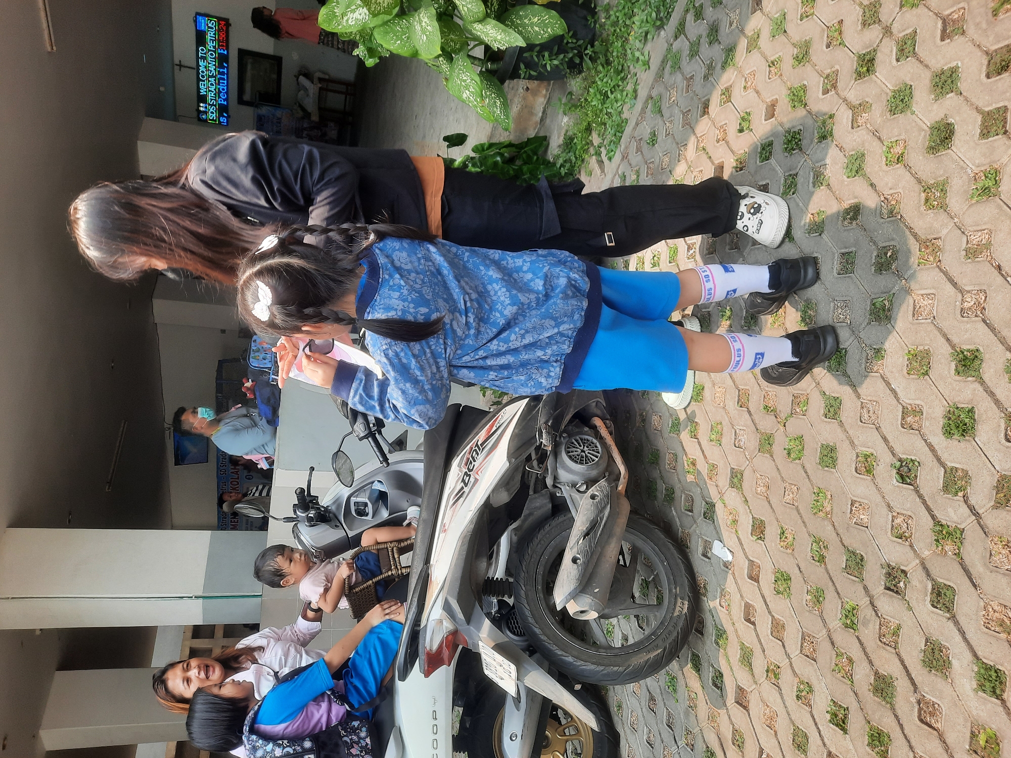 In front of a school building in North Jakarta, Indonesia, a woman and three children are gathered. A woman with brown hair and a white shirt is looking towards the left, speaking to a young girl wearing a blue shirt and backpack, who is sitting on a scooter. Next to the girl on the scooter is a toddler wearing a pink shirt, facing away from the camera. To the right of the scooter, a woman with long dark hair, wearing a black jacket and black pants, is standing with her back to the camera. She is looking down at a young girl in a blue jacket and blue shorts who is holding a piece of paper up to her face. The ground is paved with interlocking concrete blocks with grass growing in the gaps. In the background, there is a sign that reads "WELCOME TO SDS STRADA SANTO PETRUS". A motorcycle is parked behind the children and scooter.