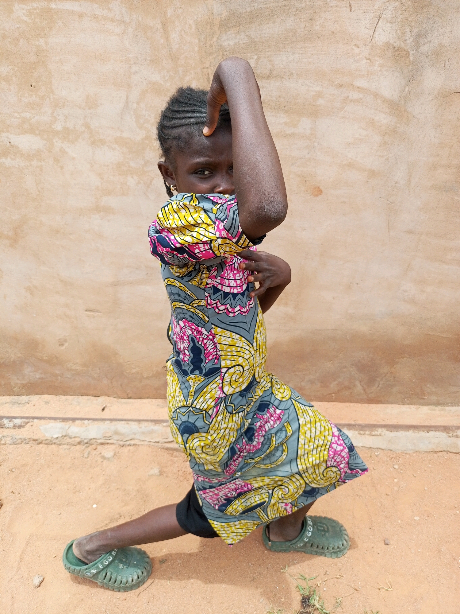 A young girl stands in a playful pose against a textured, light brown wall. She is wearing a brightly colored dress with a vibrant pattern of yellow, pink, and blue swirls and shapes. Her left arm is raised, with her hand touching her forehead, partially obscuring her face. She is looking directly at the camera with a serious expression. Her right hand is placed on her hip. The girl is wearing dark shorts underneath her dress and green clogs on her feet. The ground is sandy and reddish-brown, with a few small pebbles and sparse green weeds. The wall behind her appears to be made of adobe or stucco, showing some signs of age and texture. The lighting suggests it is daytime, likely outdoors. The overall impression is of a candid moment, capturing the girl's playful spirit and individuality in a simple, natural setting in Tempure, Nigeria.