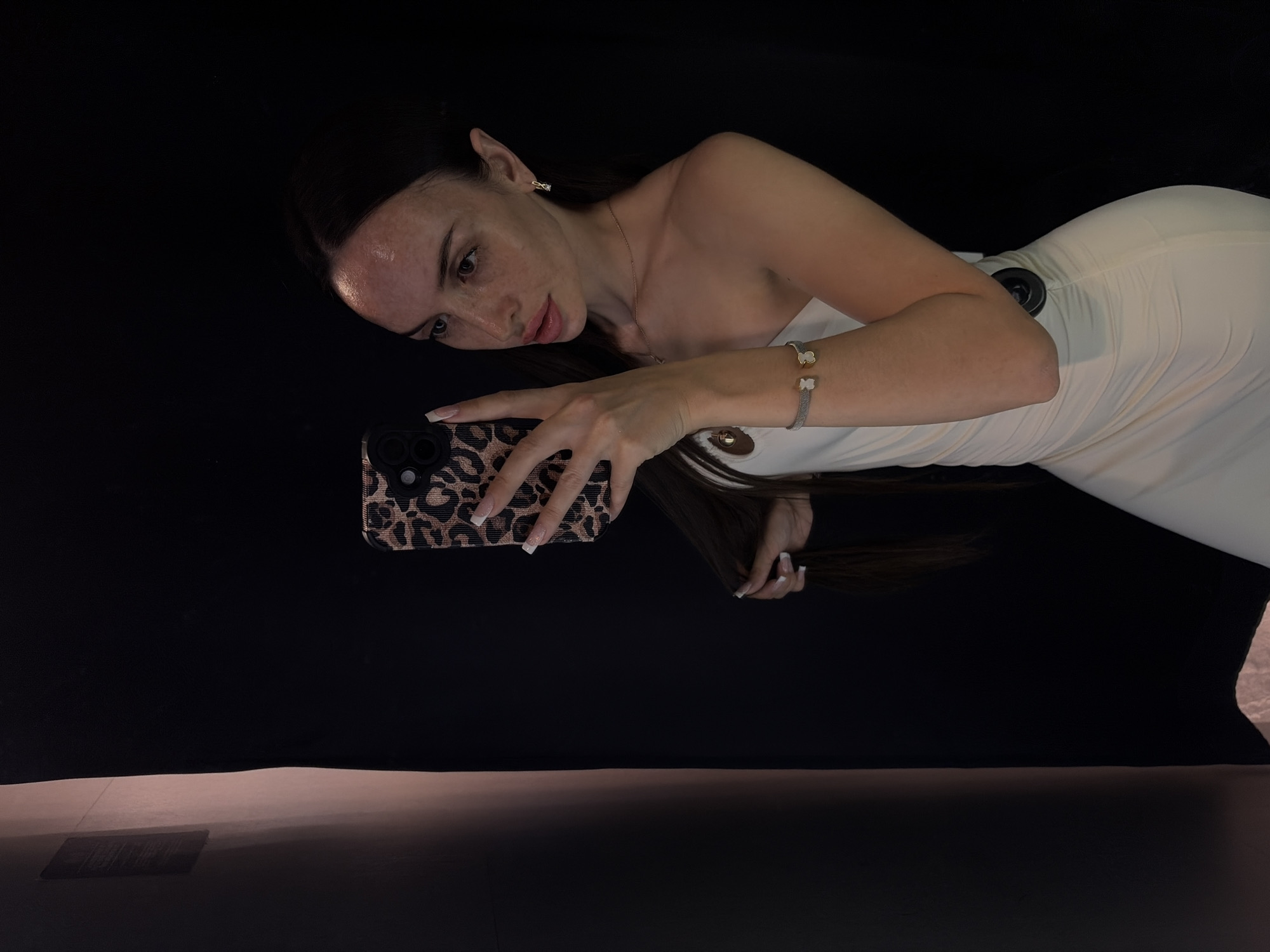A woman is taking a selfie in a dimly lit room. She is wearing a white, strapless, ruched dress with a black circular detail on the side. She has long, dark hair, and is wearing a bracelet and earrings. She is holding a phone with a leopard print case. The background is dark, with a pinkish-brown textured surface on the left. The scene appears to be indoors, possibly in a fitting room or a studio. There is no discernible information within the image to suggest a specific location in Saint Petersburg, Russia.