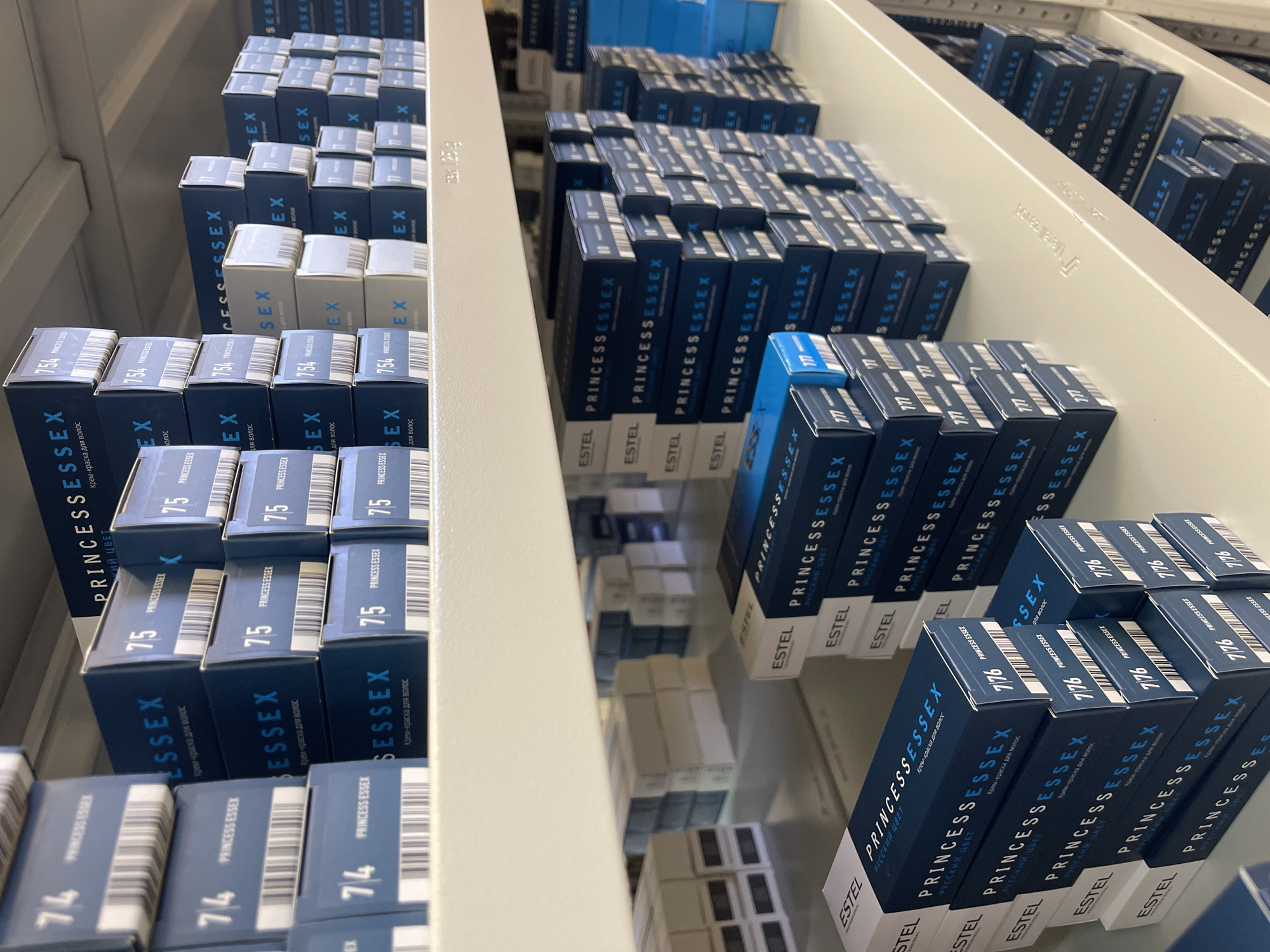 The image shows several open drawers in a shelving unit, likely located in a retail or storage area. Each drawer is filled with blue and white rectangular boxes of "Princess Essex" hair dye by Estel. The product's Russian name "РУССКИЙ ЦВЕТ" and the phrase "крем-краска для волос" (hair dye cream) are visible on the boxes.
The boxes are stacked in rows, revealing various shades numbered 74, 75, 754, 777, and 7/76. The shelves appear to be light gray, with some text and numbers visible on the drawer edges. The photograph's perspective suggests it was taken from a slightly elevated position. The location mentioned, Chaikovsky, Russia, cannot be verified from the image alone but is contextually relevant given the Russian text on the product boxes. There are no people visible in the image, and the focus is solely on the stocked hair dye products.