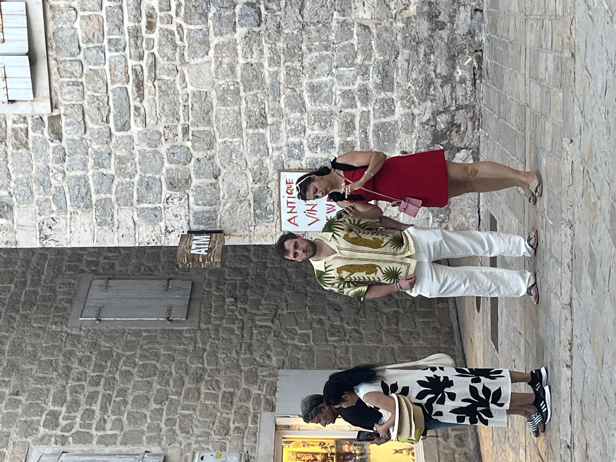 In Budva, Montenegro, four individuals are observed in the foreground of a weathered stone building. On the right, a man with dark hair wears a short-sleeved, patterned shirt featuring pineapples and leopards, paired with white trousers and flip-flops. Beside him, a woman with dark hair tied back, wearing a red dress and white sunglasses on her head, holds a pink handbag and a smartphone. To the left, a woman in a black and white floral dress stands next to a man with graying hair and sunglasses, who is holding a smartphone. The background reveals stone walls and a sign that reads "ATM." The ground is paved with cobblestones.