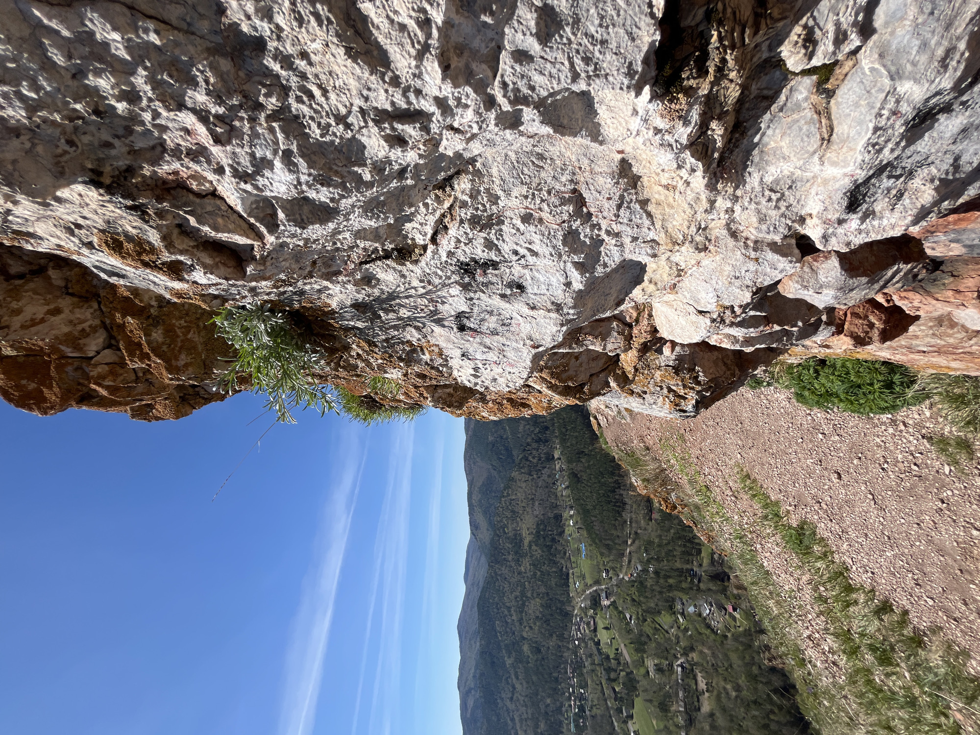 The image depicts a view from a rocky precipice overlooking a mountainous landscape with a town nestled in the valley. The foreground is dominated by the rocky cliff face, showing various textures and coloration, from gray to brown. Small plants are growing from crevices in the rock. Below the cliff, there is a steep slope covered in loose rock and vegetation, leading down to the valley. The valley contains a town with buildings and roads visible amongst the trees. In the distance, there are mountains and a blue sky with thin clouds. The location is likely in or near Krasnoyarsk, Russia, based on the landscape and geographic features. There are no people visible in the image.