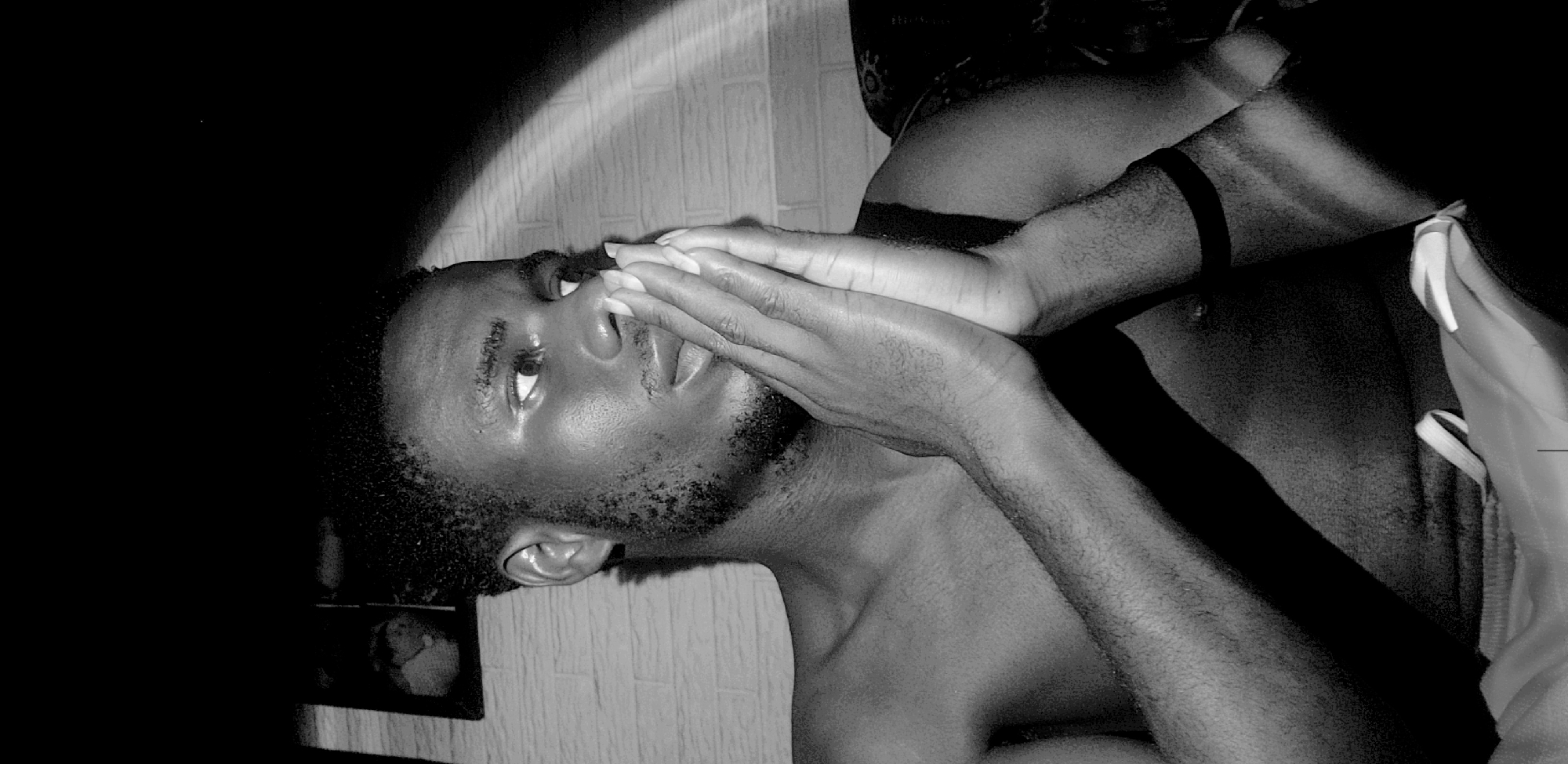 This high-contrast black and white medium shot captures a young man indoors, likely in Narayi, Nigeria. The man, with short, dark hair and a beard, is shirtless and appears to be lying down, looking upwards with an earnest expression. His hands are clasped together, partially covering his nose and mouth in a gesture that suggests contemplation, prayer, or deep thought.
The scene is dramatically lit by a strong, arched beam of light that illuminates his face, chest, and a portion of the textured wall behind him, creating sharp highlights and deep shadows across the image. The rest of the space is largely obscured by darkness. A black wristband is visible on his right arm. In the bottom left corner, a small, framed picture rests on a surface, faintly visible amidst the shadows. On the right side, there's a slight glimpse of light-colored fabric or clothing. The strong play of light and shadow emphasizes the man's pensive mood and the intimate setting. No text is visible in the image.