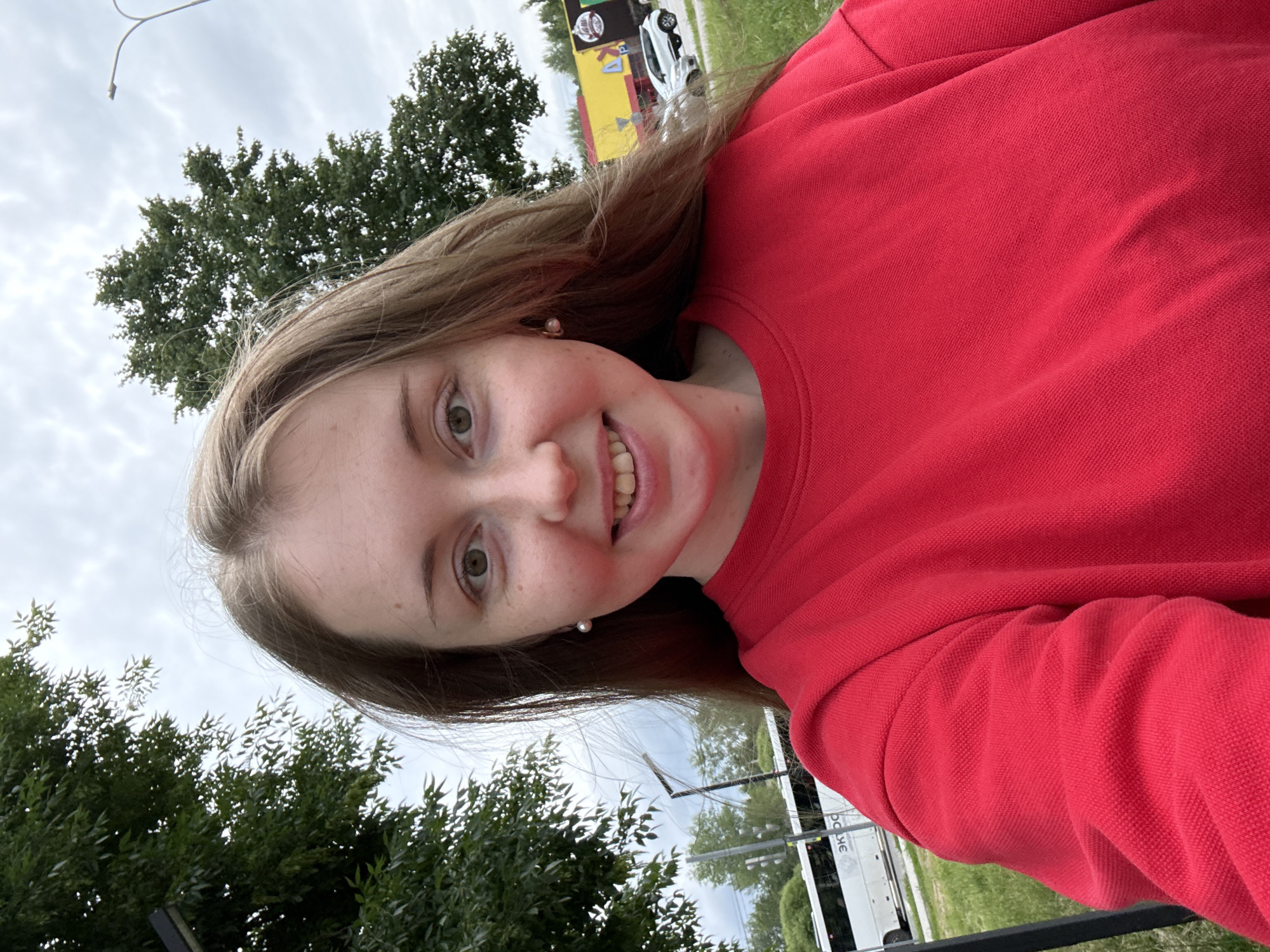 A young woman with long, light brown hair and green eyes is shown in a selfie outdoors. She is wearing a bright red long-sleeved shirt and small pearl earrings. She is smiling, revealing some of her teeth. In the background, there are trees, a cloudy sky, a white bus with Cyrillic lettering, a lamppost, and a building with yellow and dark brown sections. The setting appears to be a street or road with some greenery.