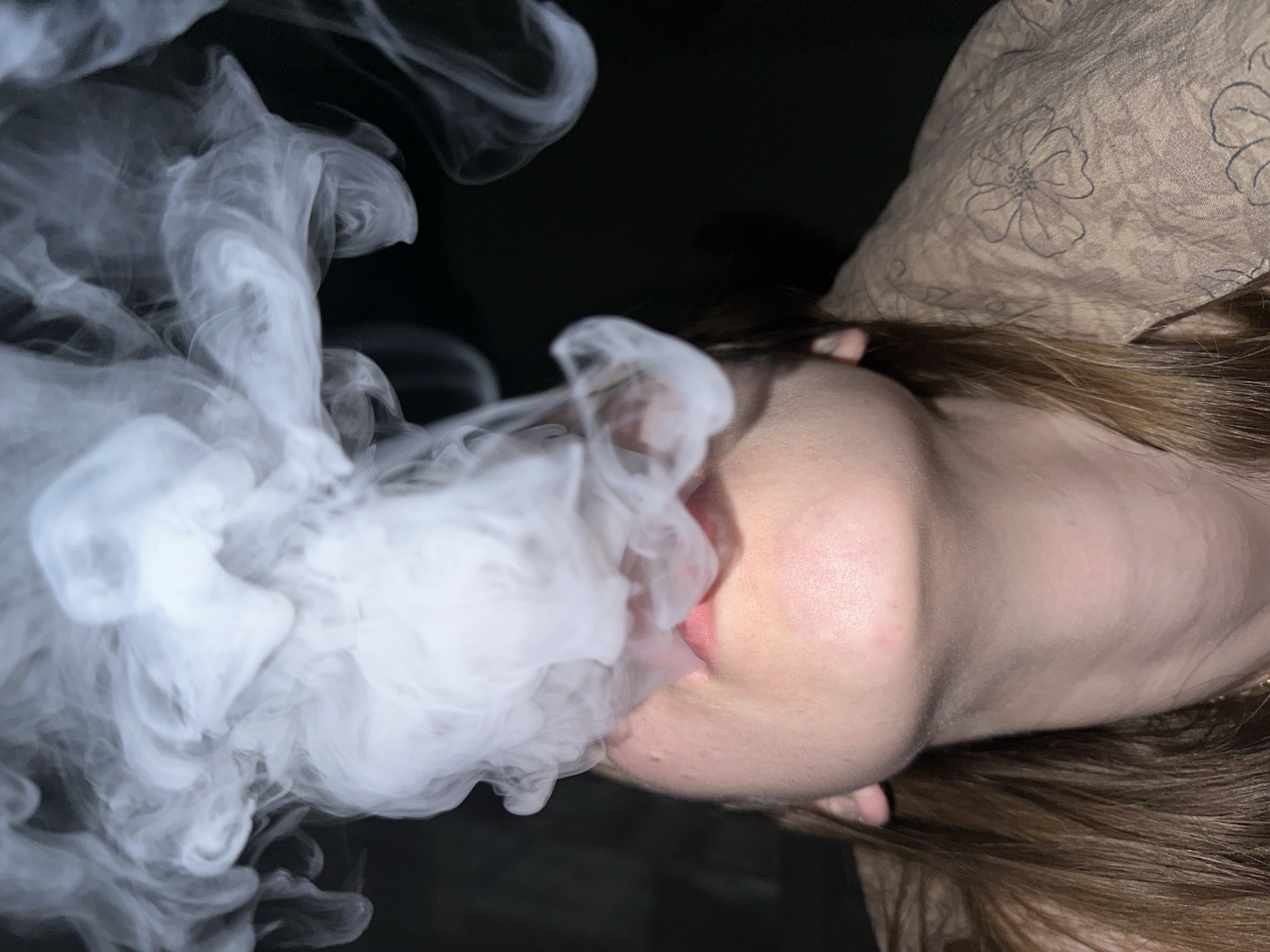 This image captures a close-up, low-angle view of an individual's lower face, neck, and upper shoulder, located in the city of Chaikovsky, Russia. The subject, exhibiting fair skin and long brown hair visible on the left, is actively exhaling a dense, white cloud of vapor or smoke, which billows upwards from their slightly parted lips. The person is wearing a light brown or beige garment patterned with dark floral outlines, visible on the right shoulder. A thin gold chain is present around the neck, and a small gold earring is visible in the left earlobe. The background is uniformly dark, providing no additional environmental context.