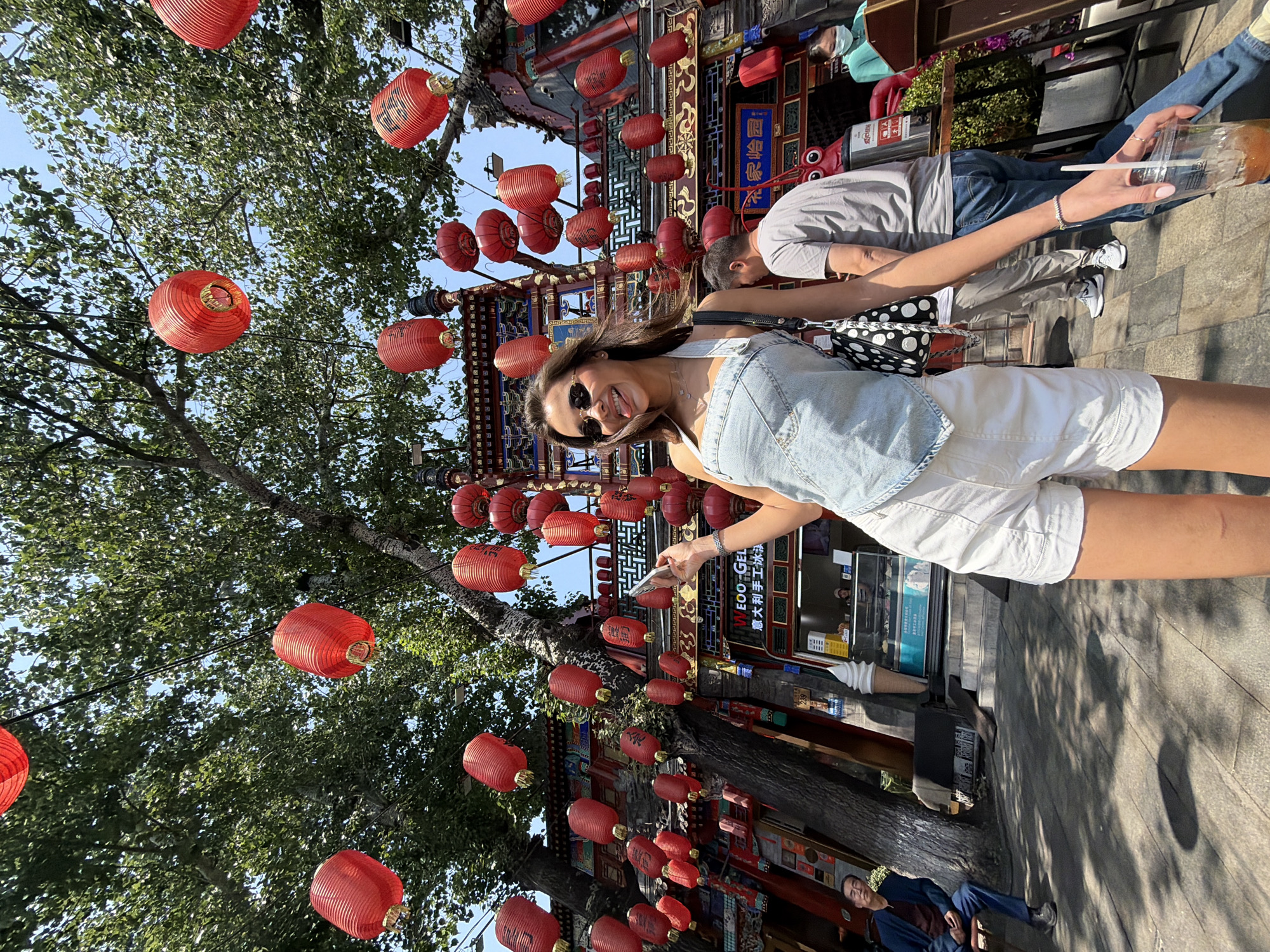 A medium, eye-level outdoor shot depicts a woman holding a phone on a sidewalk lined with trees and red lanterns in the Dongcheng District of Beijing, China.
The woman, centered and facing the camera, wears sunglasses and a light-colored denim jacket over a white outfit. She holds a phone in her left hand. Beside her, on the right, an unidentified person holds a drink.
Red Chinese lanterns are strung above, interspersed among the trees. In the background are buildings featuring traditional Chinese architectural elements. The sidewalk is concrete, and a person sitting on a bench can be seen in the lower right corner.