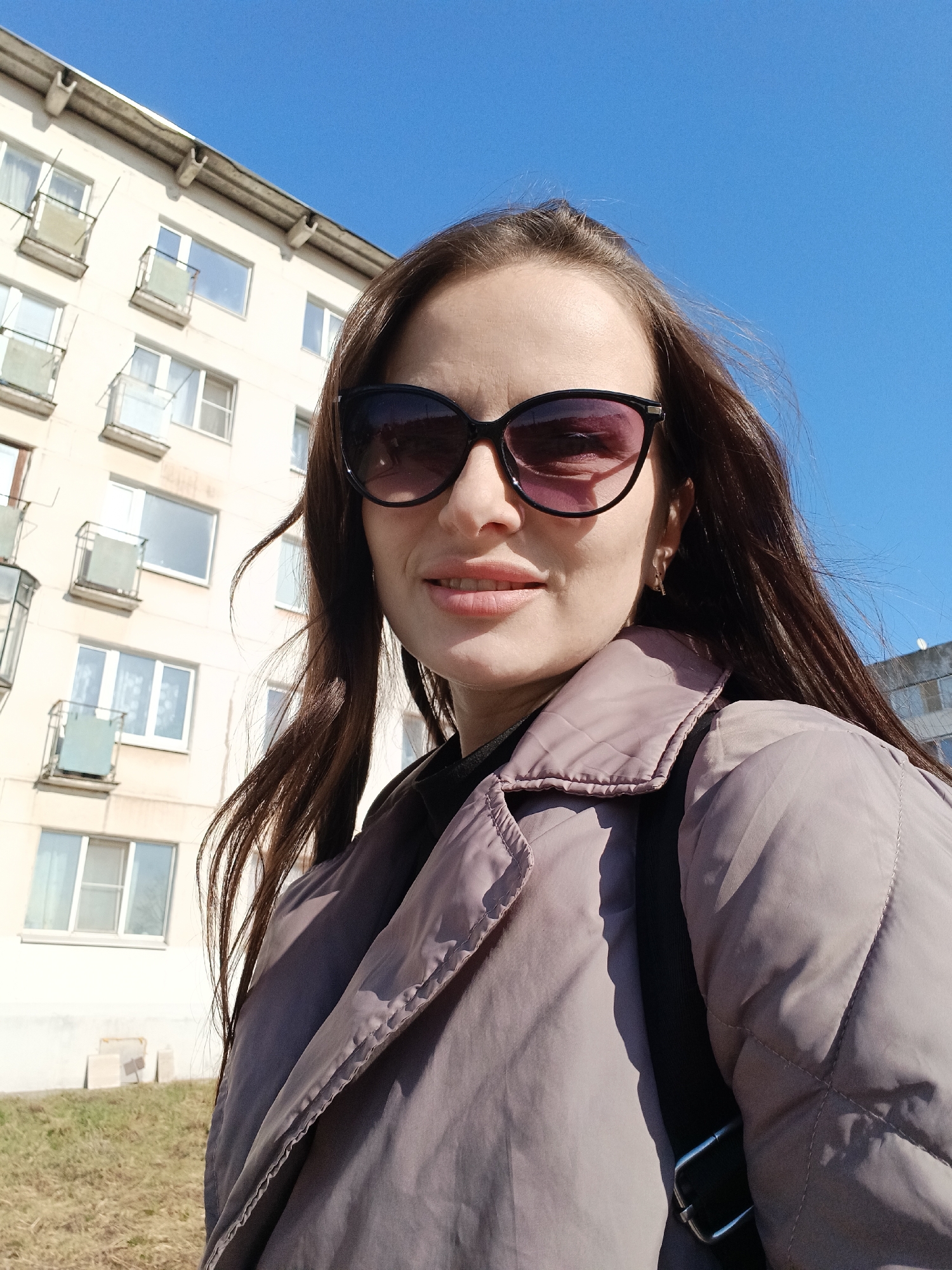 A woman in a quilted jacket and sunglasses is posing for a selfie on a sunny day in an urban setting. The background shows a multi-story apartment building with balconies under a clear blue sky.