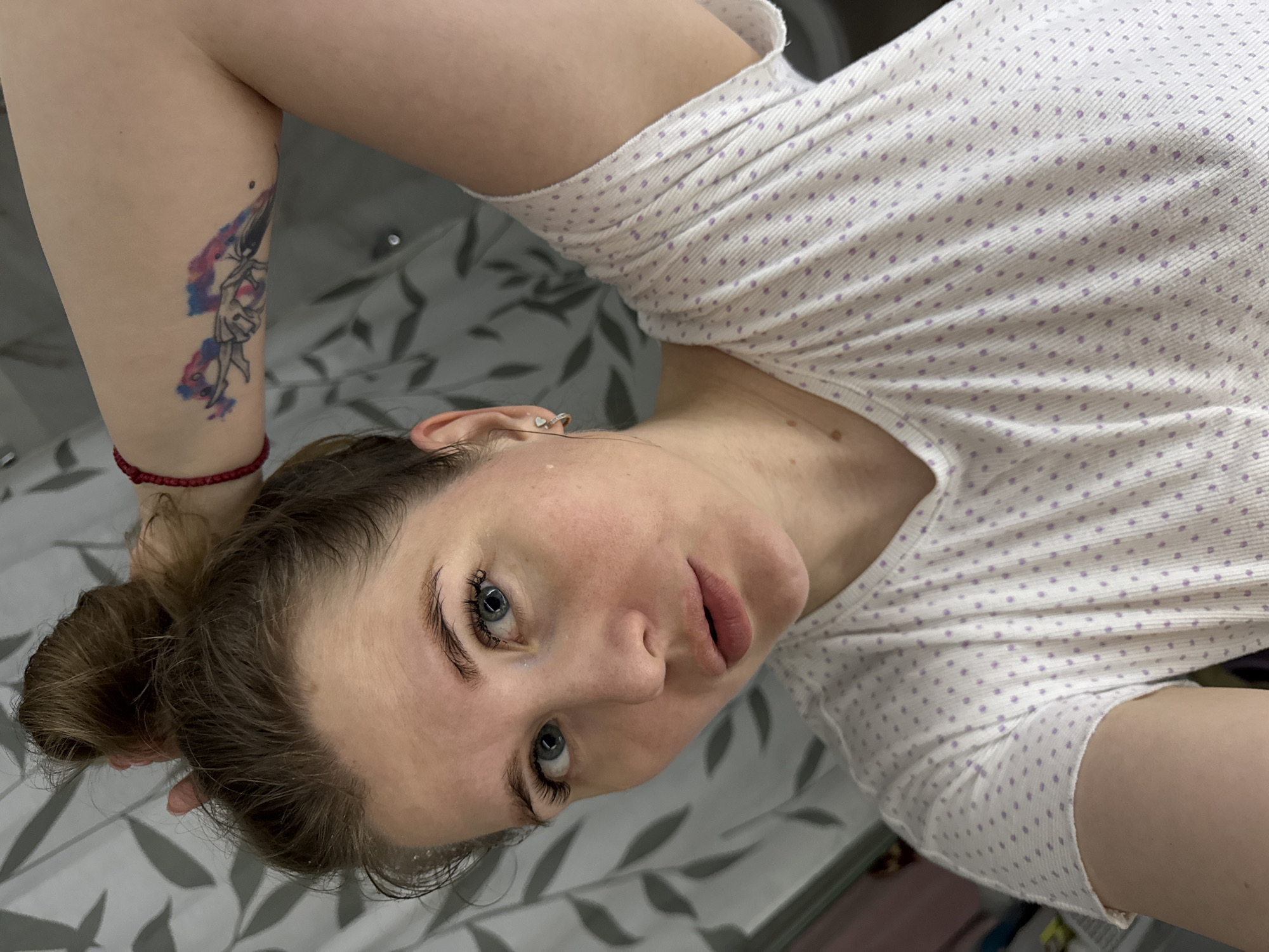 A fair-skinned woman with blue eyes and light brown hair pulled back in a bun lies on a bed with a grey and white leaf-patterned sheet. She wears a white t-shirt with small purple polka dots. She has a tattoo on her upper left arm consisting of a blue and purple design. A red bracelet is visible on her left wrist. She appears to be in an indoor location. The location within the city of Gornyi (Dvurechenskoe S/P), Russia, cannot be determined from the image.
